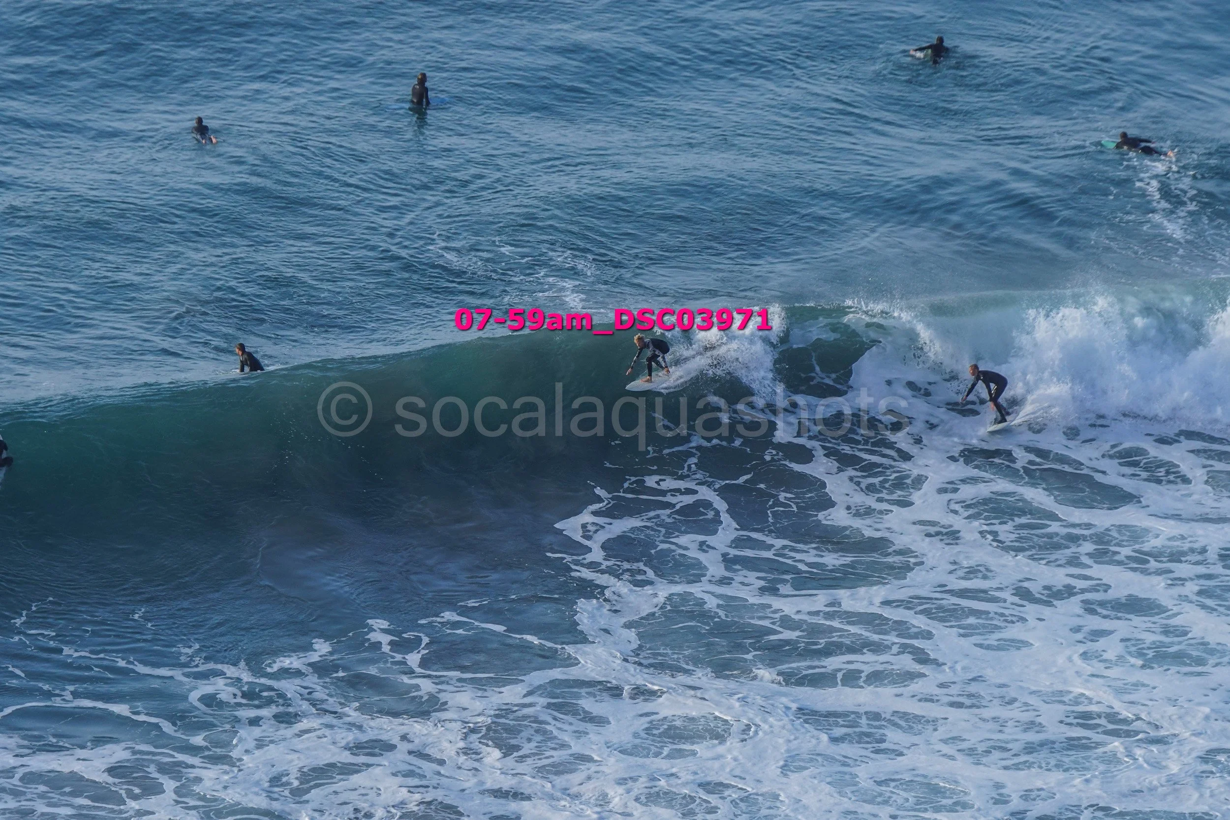 Multiple surfers riding and waiting on ocean waves, some in the water and some on the surfboard, under a cloudy sky.