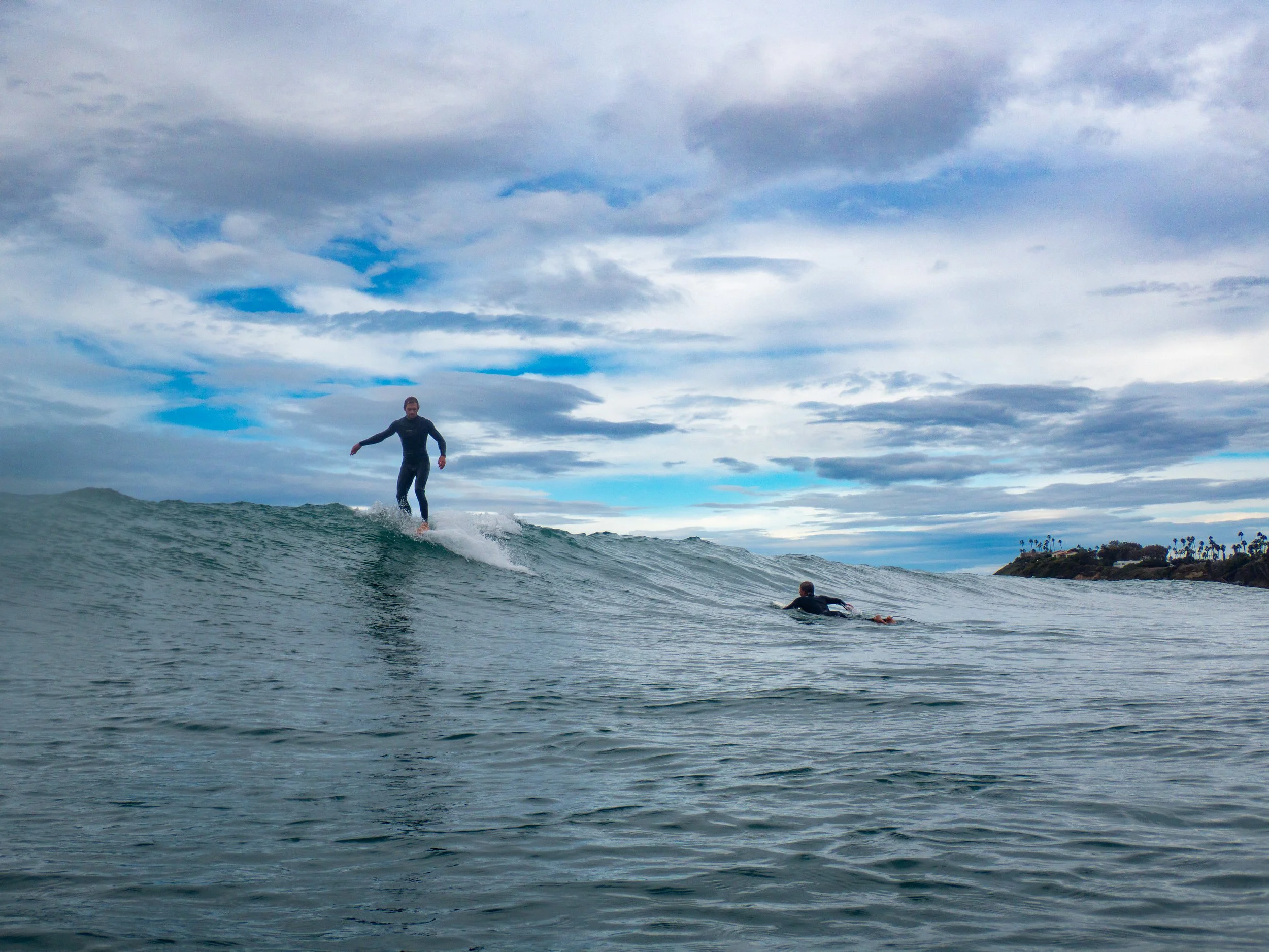 A person surfing on a wave in the ocean, with another person swimming nearby and a coastline with palm trees in the distance under a cloudy sky.