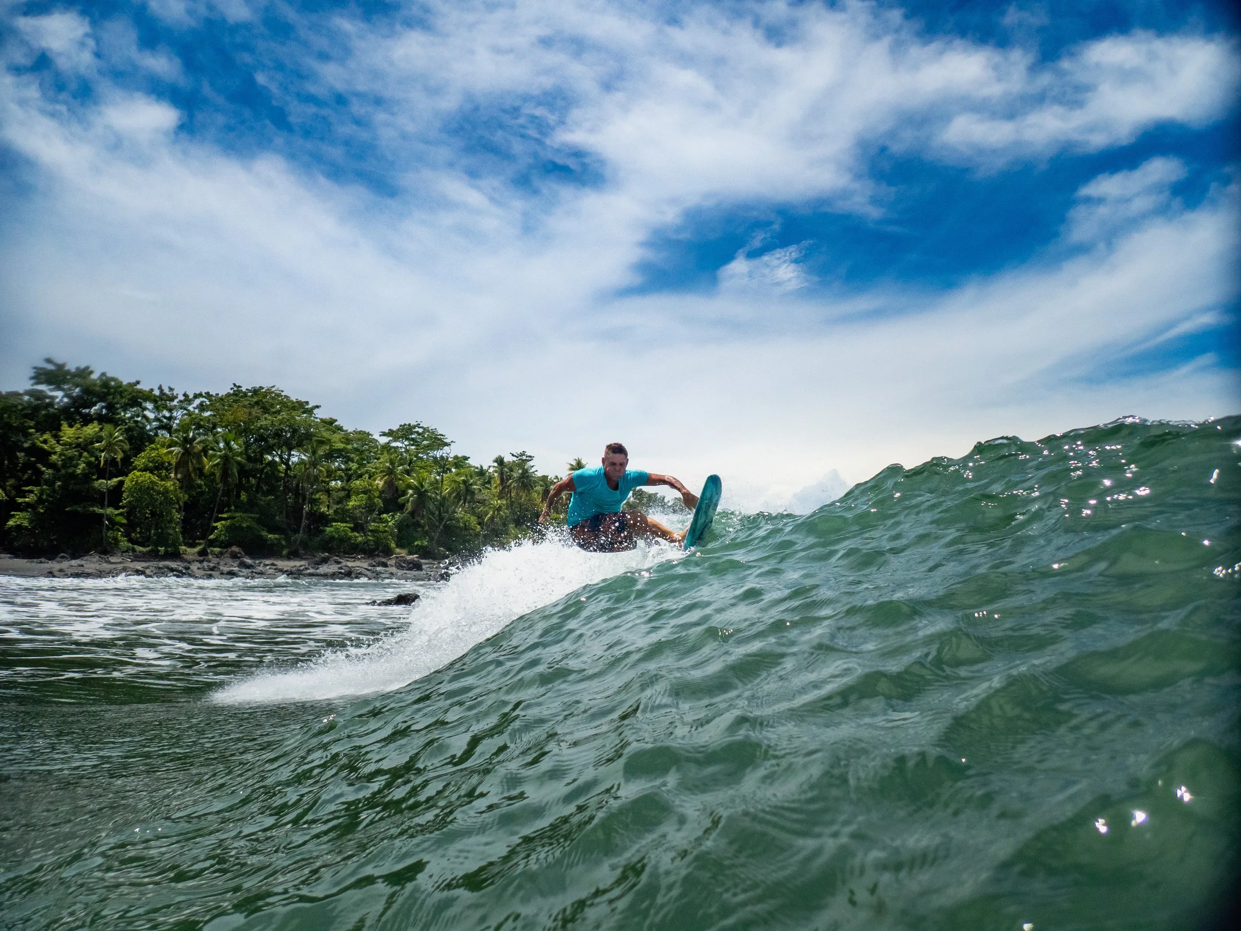 Person surfing on a wave with a tropical coastline and blue sky in the background.