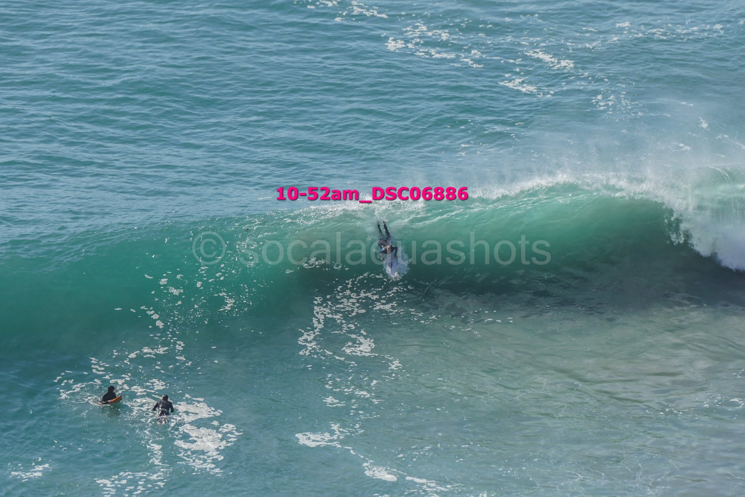 A person surfing a large wave in the ocean, with two people swimming nearby.