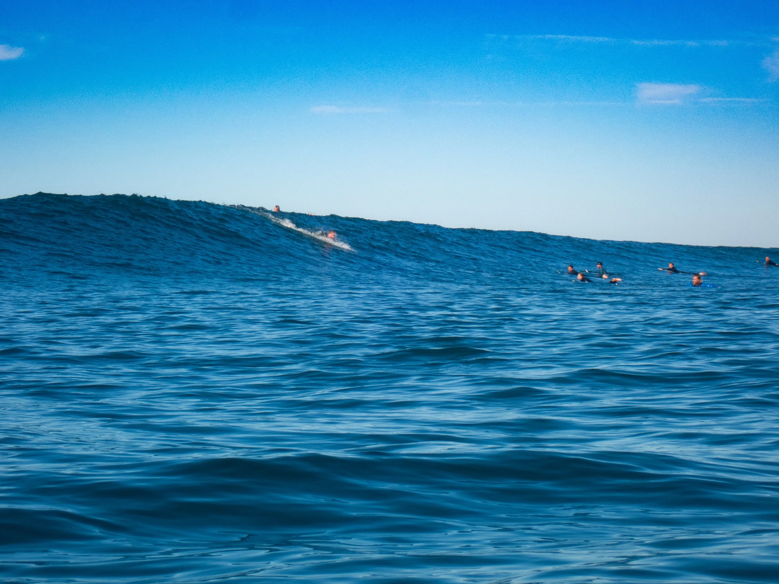 Group of people swimming in the ocean near a large wave under a clear blue sky.