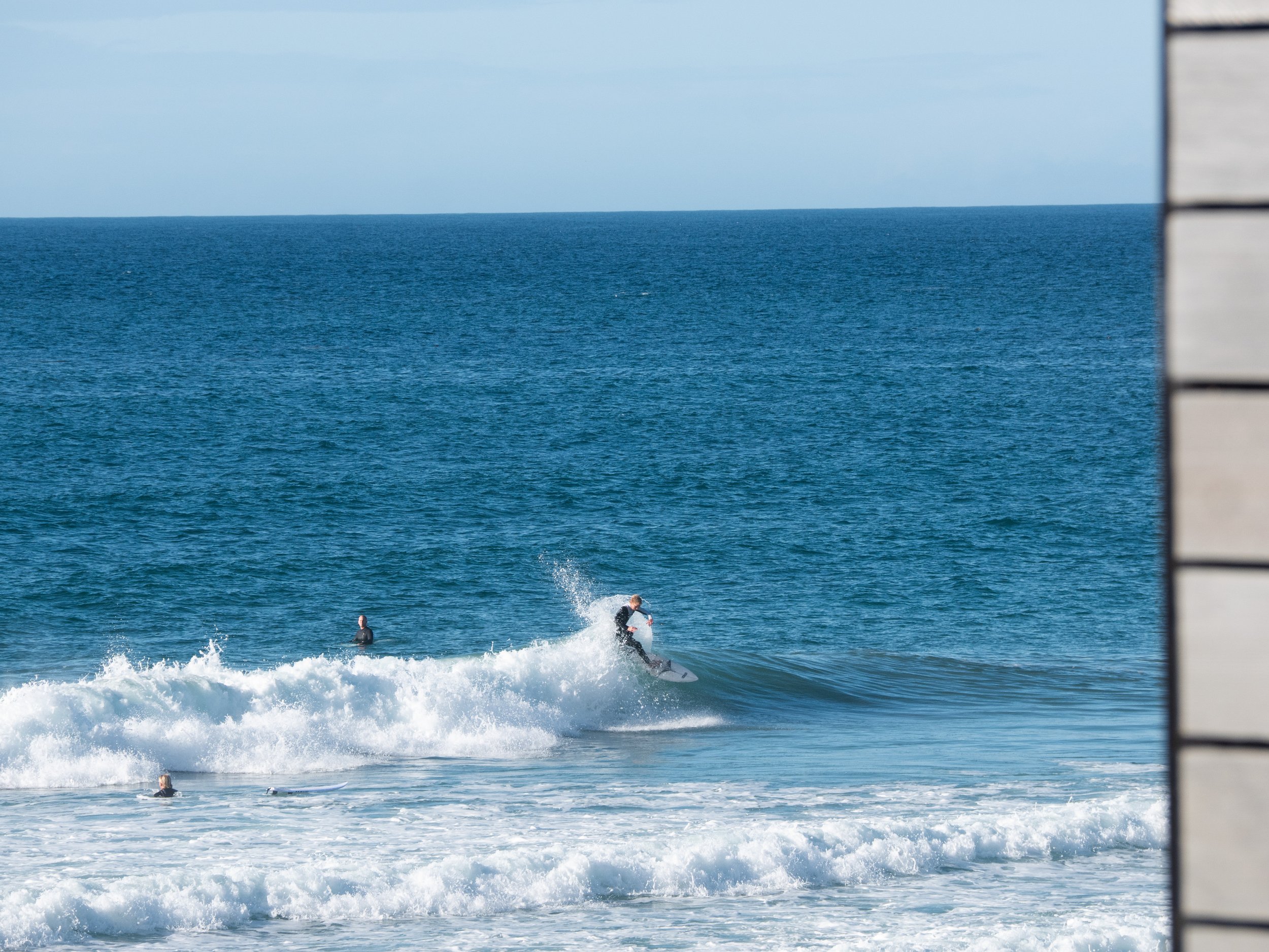 A person surfing on a wave in the ocean with a few other people in the water and a clear blue sky.