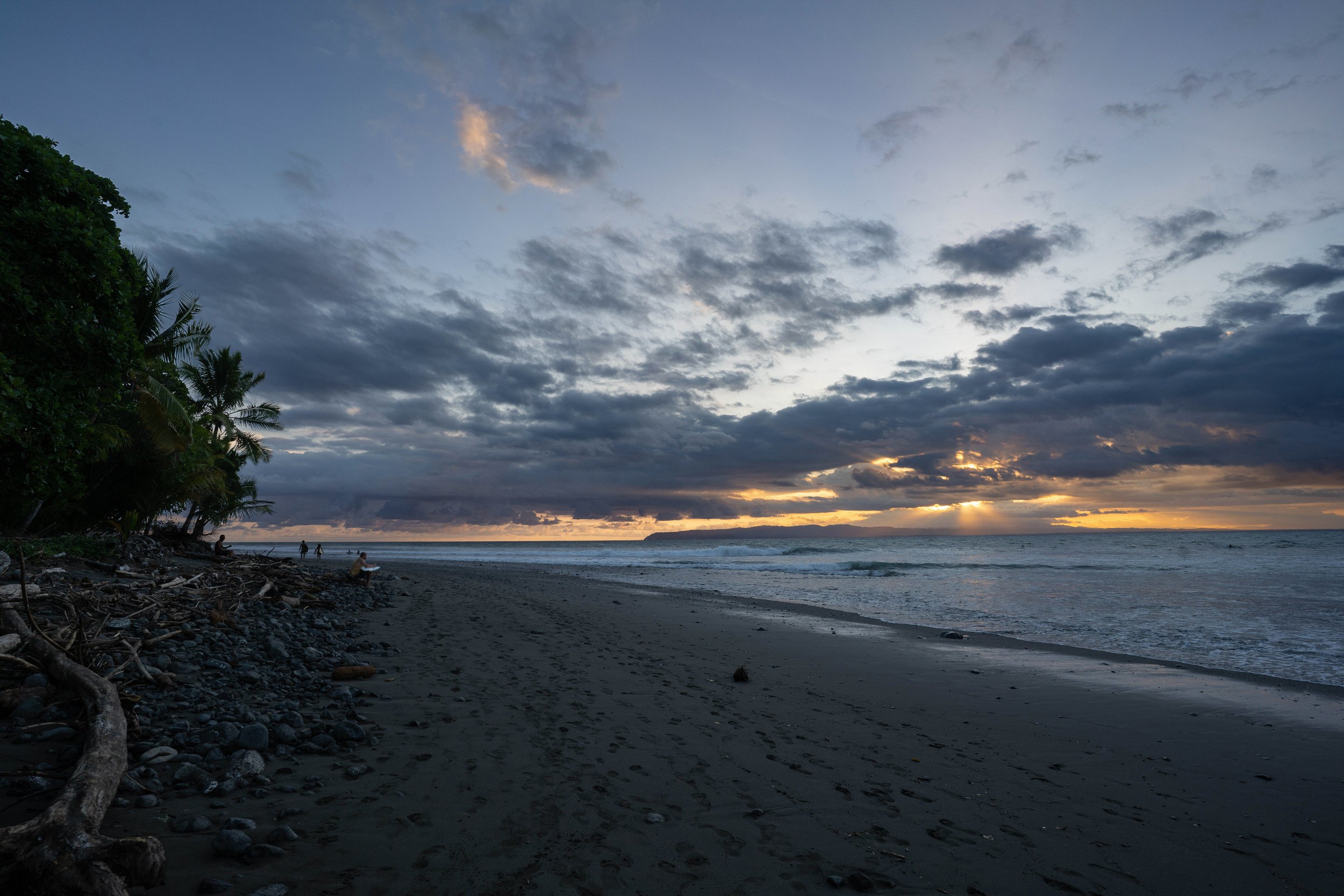 A cloudy sunset over a rocky beach with a few people walking and lush green trees on the left.