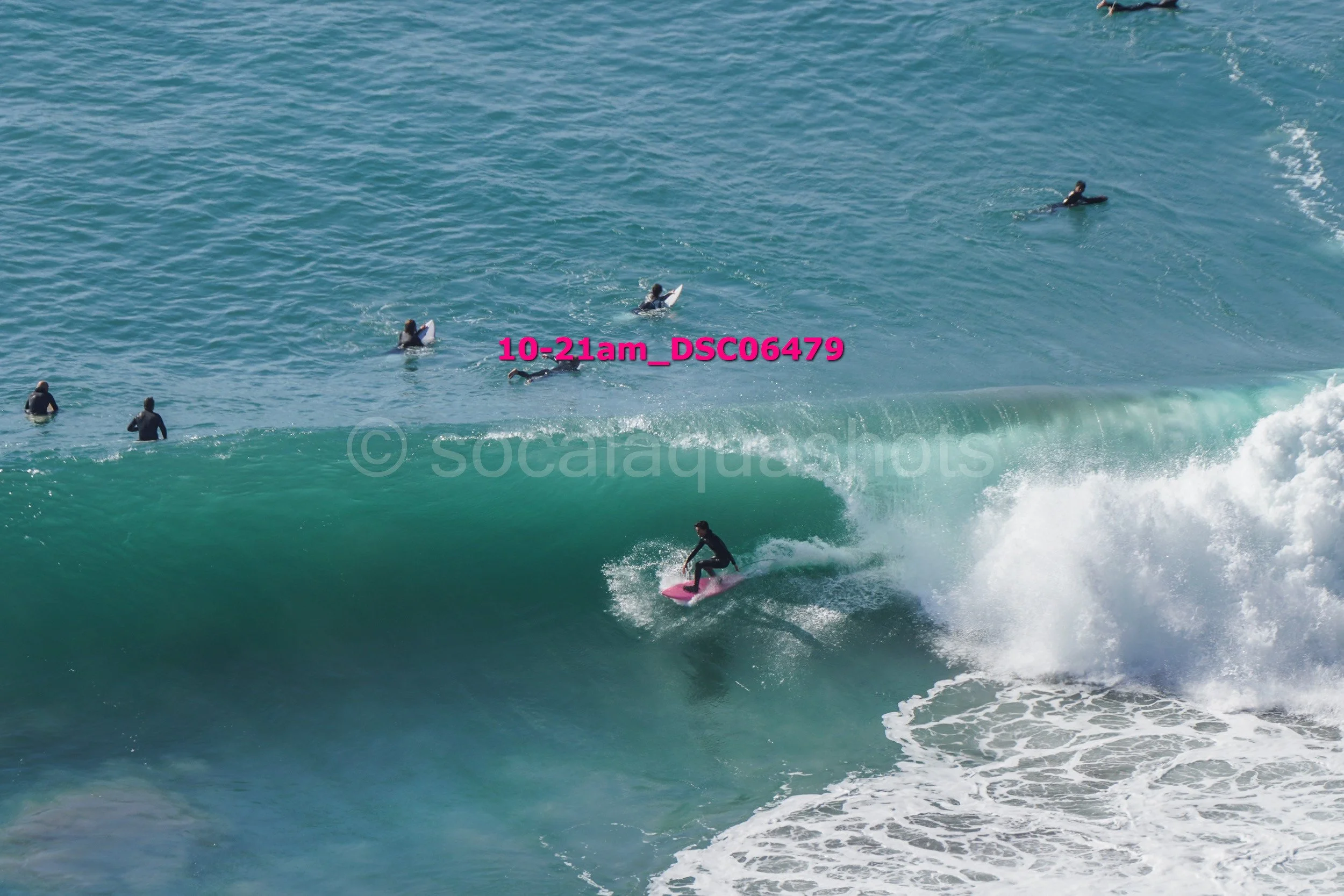 A group of surfers, some on surfboards, in the ocean with one surfer riding a wave on a pink surfboard.
