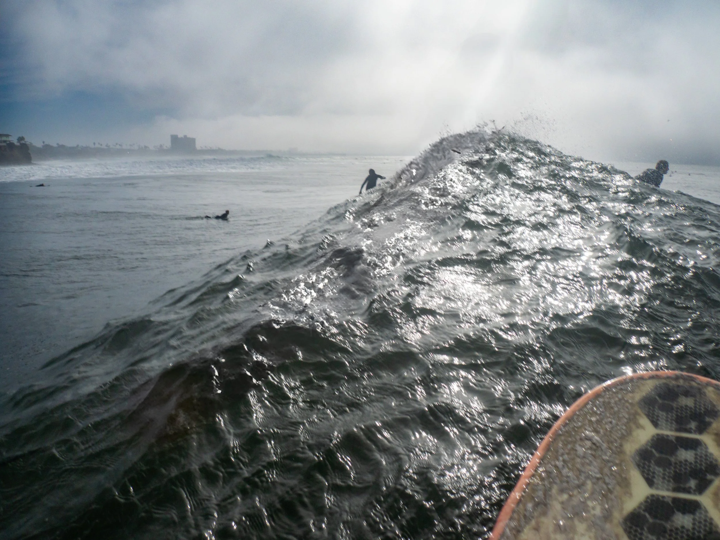 People surfing and swimming in the ocean during cloudy weather, with buildings visible on the distant coastline.