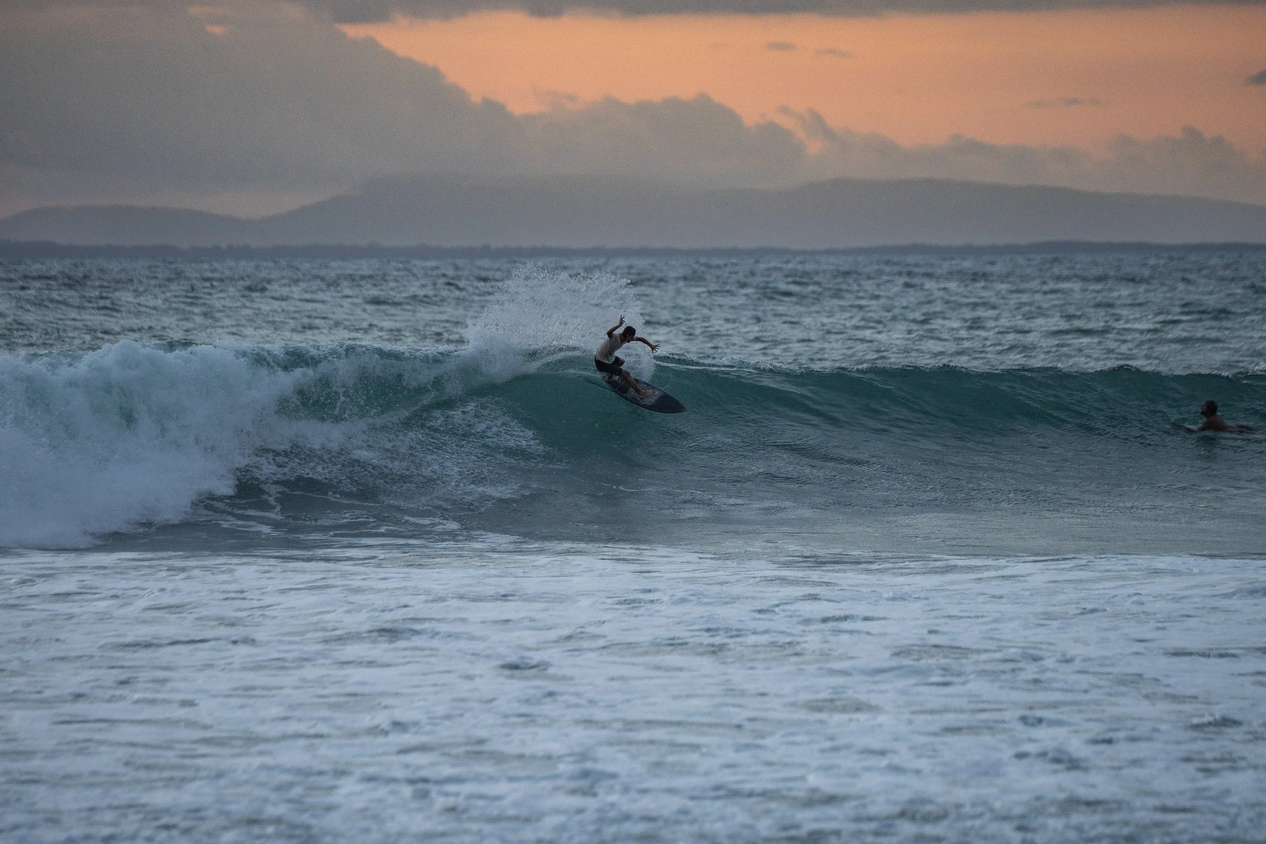 A person riding a surfboard on a wave at sunset, with another person in the water nearby and mountains in the background.