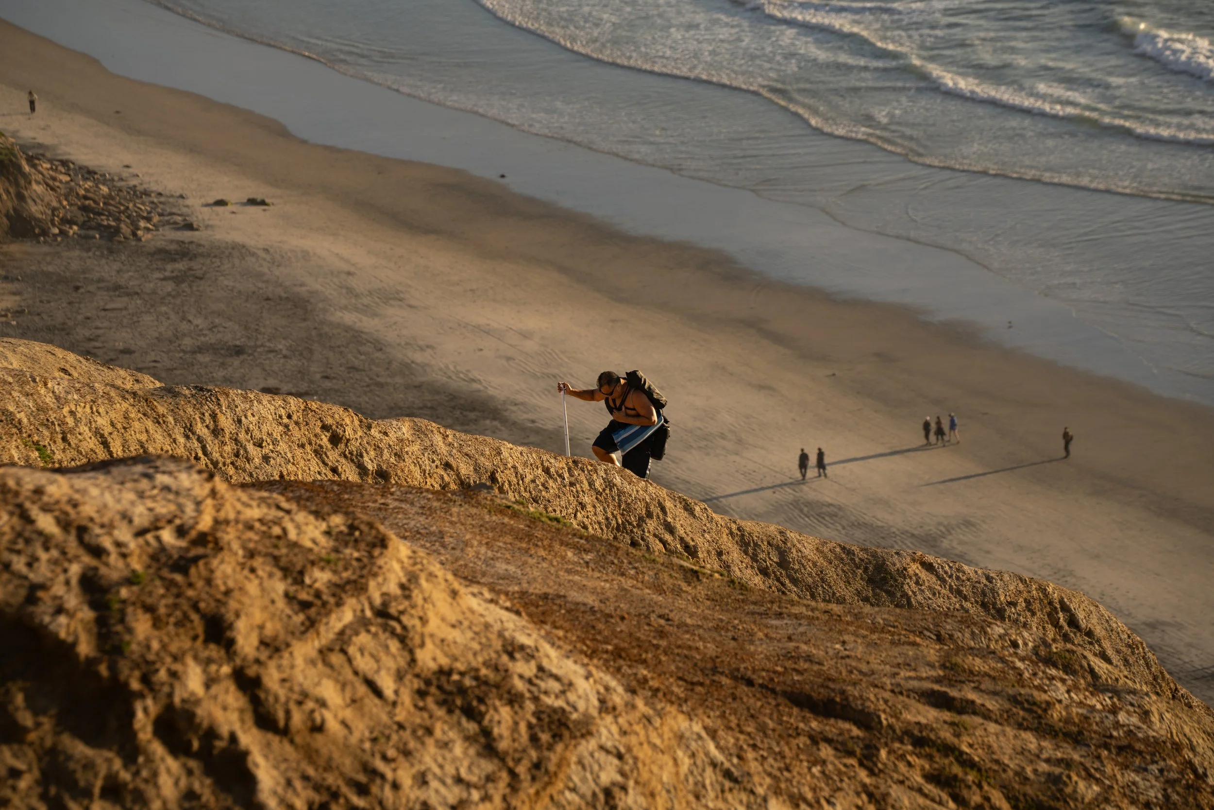 A person climbing a steep rocky hill on a beach with others walking along the sand and in the water in the background during sunset.