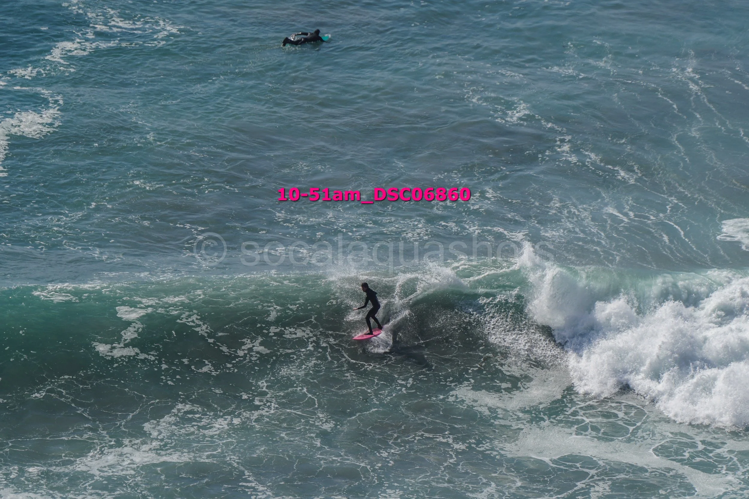 A person surfing on a pink surfboard in the ocean with a second person swimming in the water in the background.