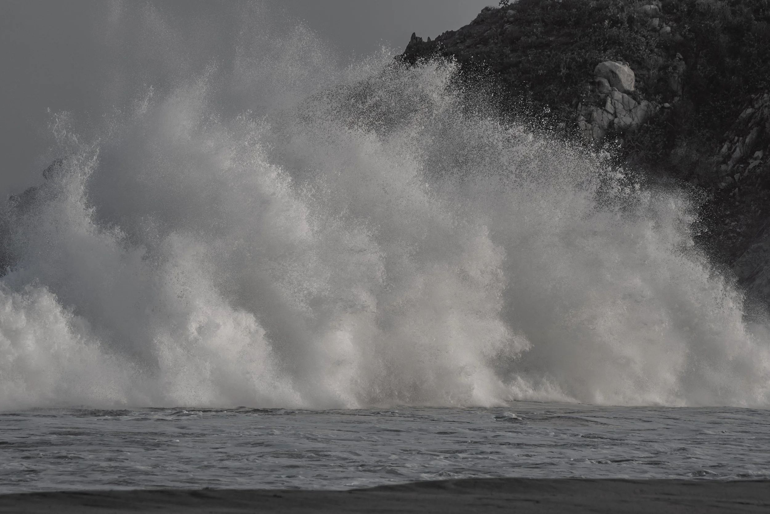 Large ocean waves crashing against rocks with spray and foam, dark gray sky.