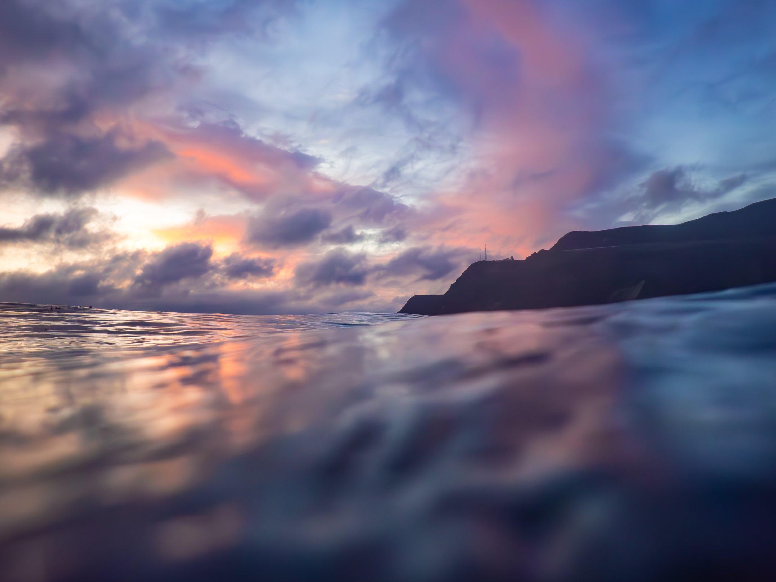 Sunset over the ocean with a silhouette of cliffs in the distance, clouds in the sky reflecting the colors of the sunset.