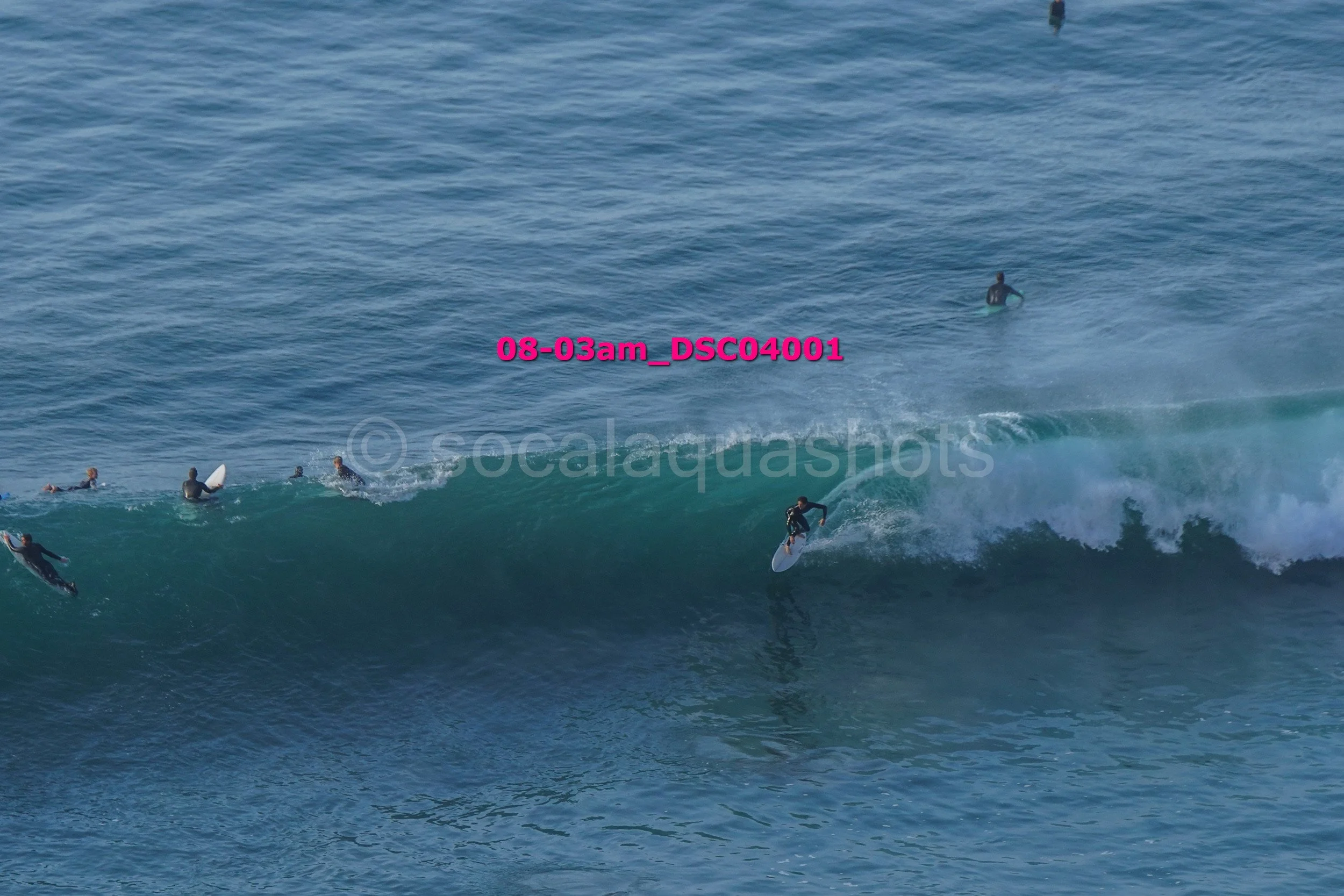 Multiple surfers in wetsuits riding and waiting on ocean waves, with some sitting on surfboards and others standing or riding the waves.