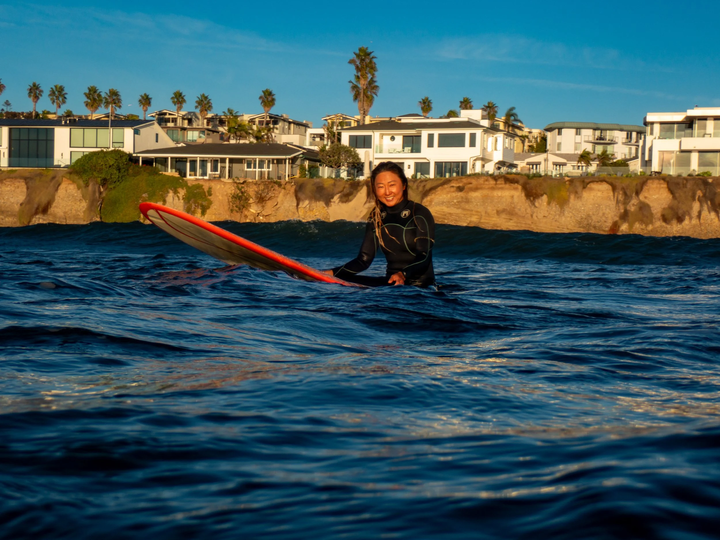 A woman in a black wetsuit is sitting on a surfboard in the ocean, smiling at the camera. There are cliffs with houses and palm trees in the background, under a clear blue sky.