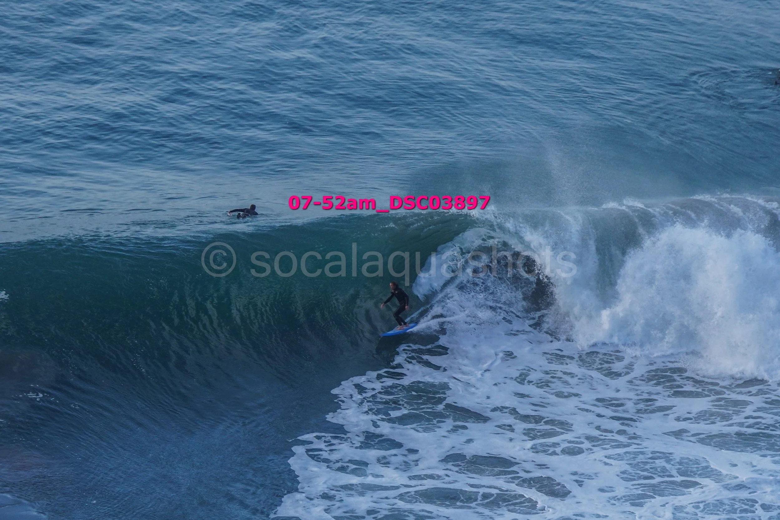 A person surfing on a large wave in the ocean, with another person swimming nearby in the water.