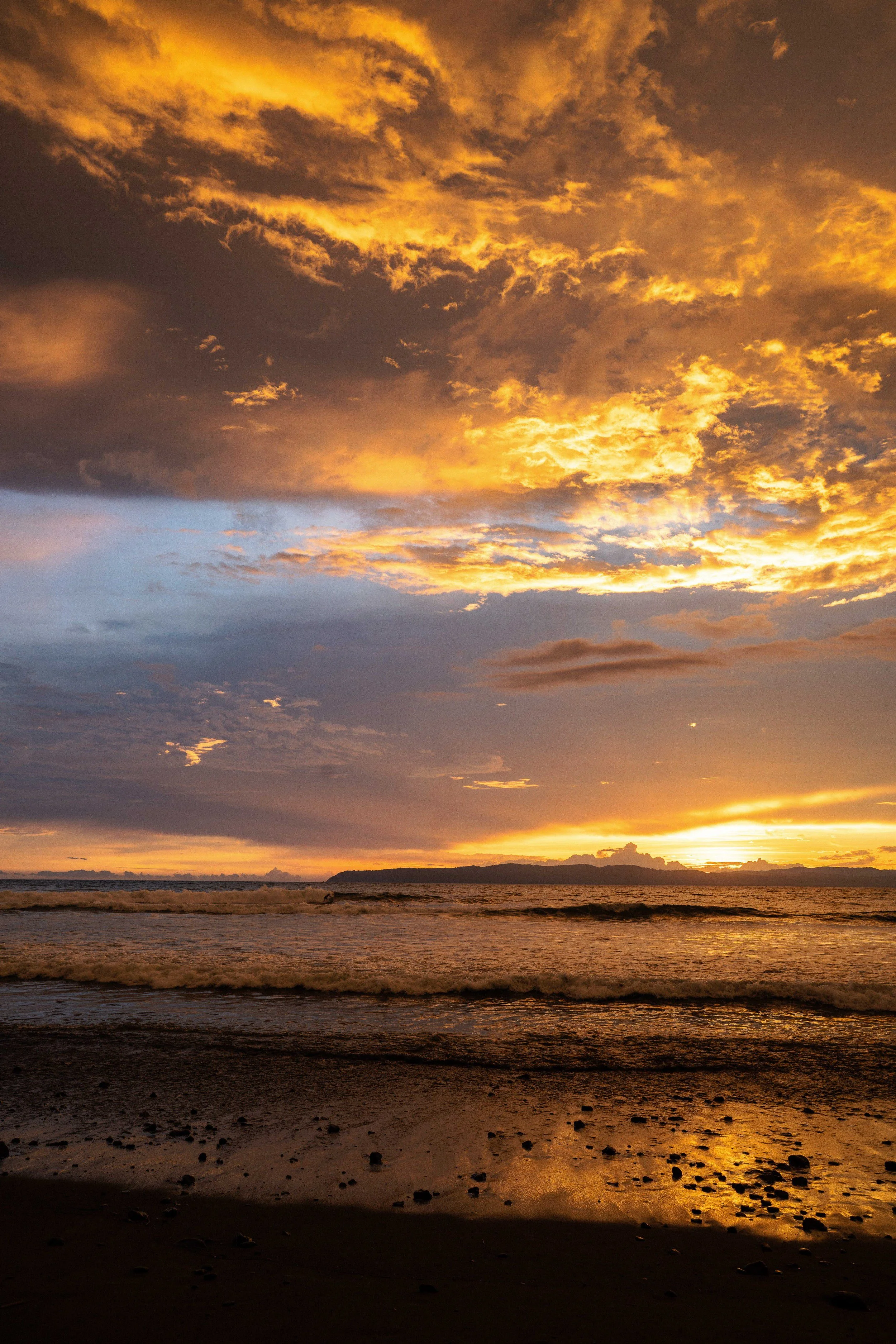 Sunset over the ocean with a sky filled with clouds, casting an orange and yellow glow, and waves gently crashing on the sandy beach.