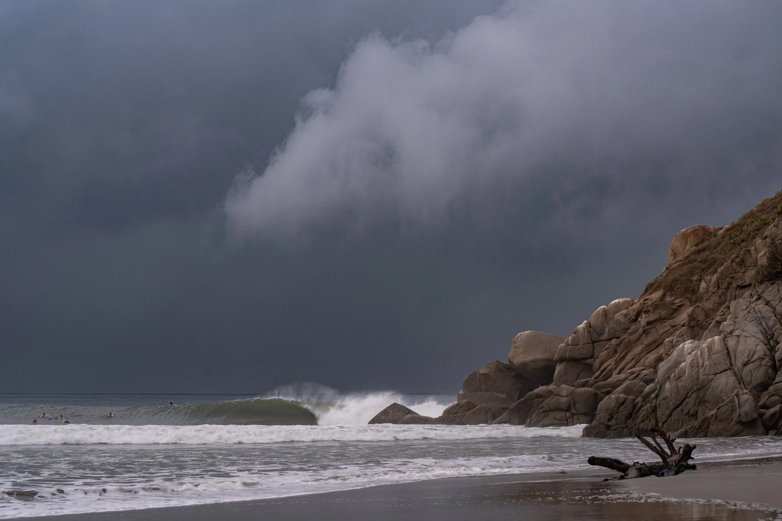 Stormy sky over a rocky shoreline with ocean waves crashing against the rocks and a few surfers in the water.