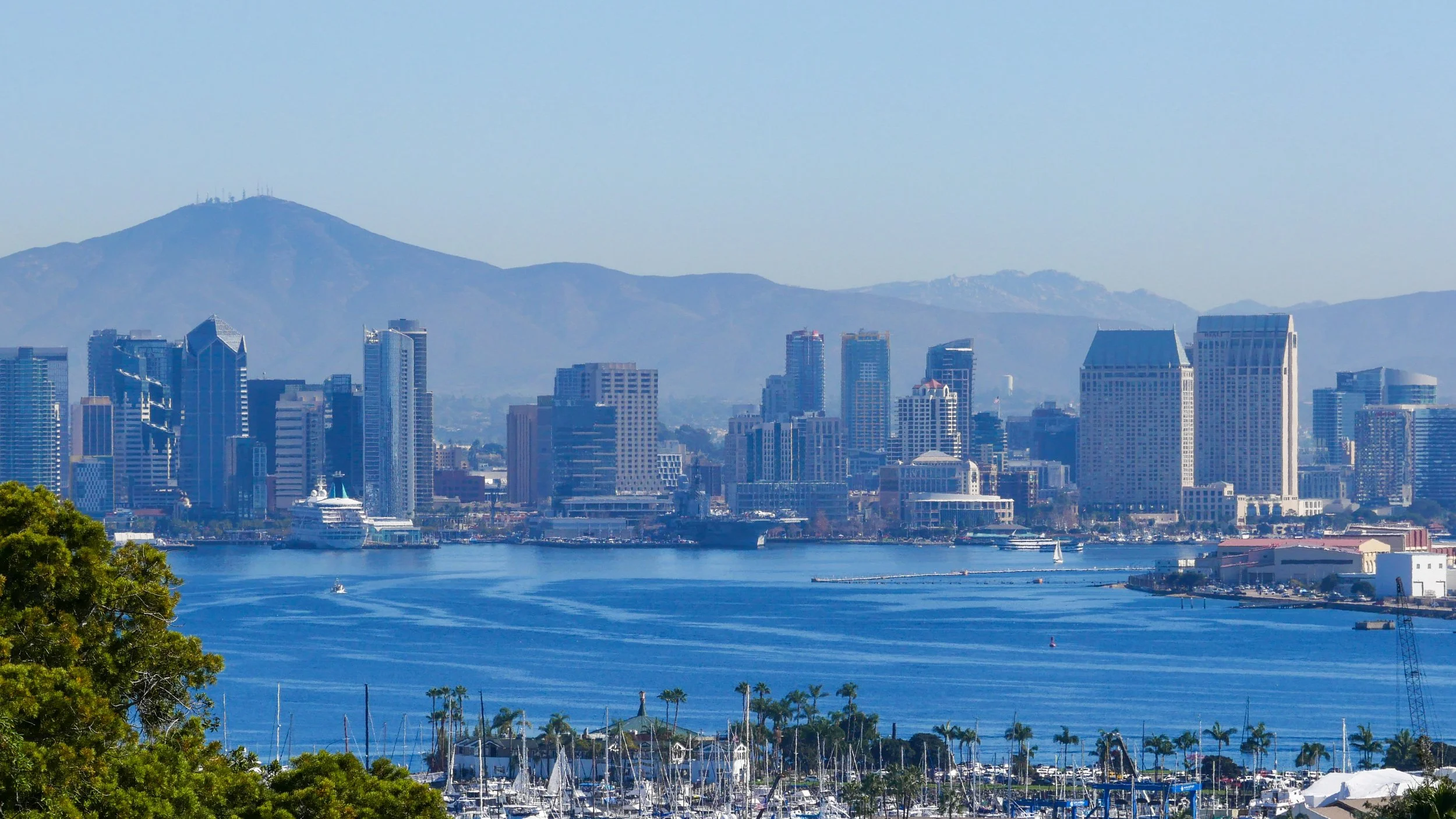 San Diego skyline with tall buildings, mountains in the background, and water with boats in the foreground.
