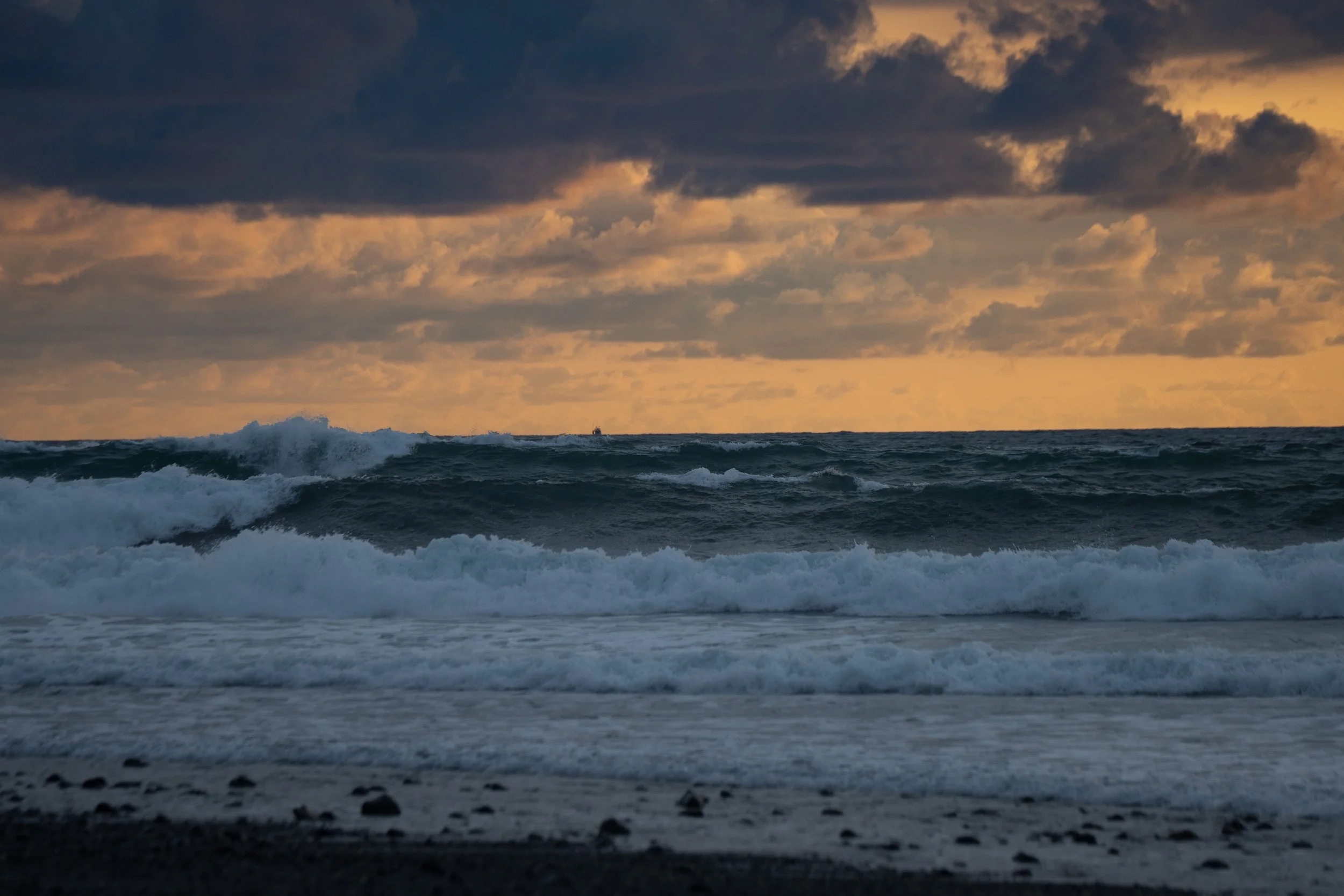 Ocean waves crashing onto the shoreline at sunset, with dark clouds in the sky and a small boat in the distance.