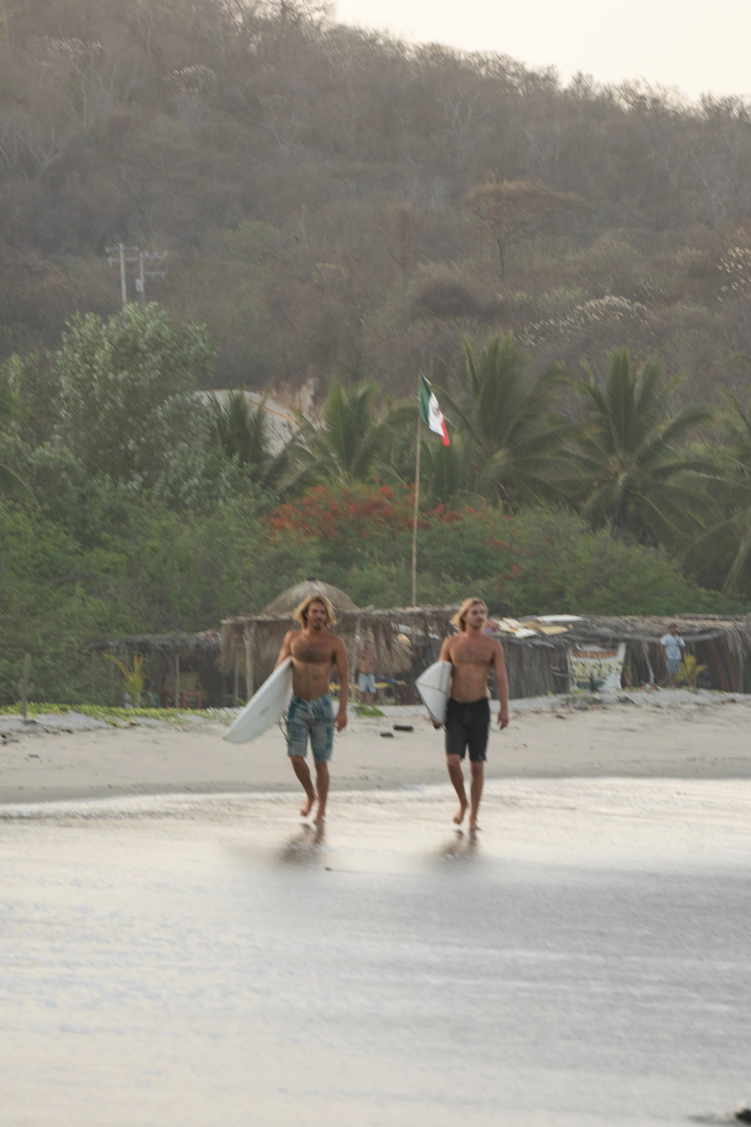 Two shirtless men walking along a beach holding surfboards, with a lush, hilly background and a Mexican flag on a pole.