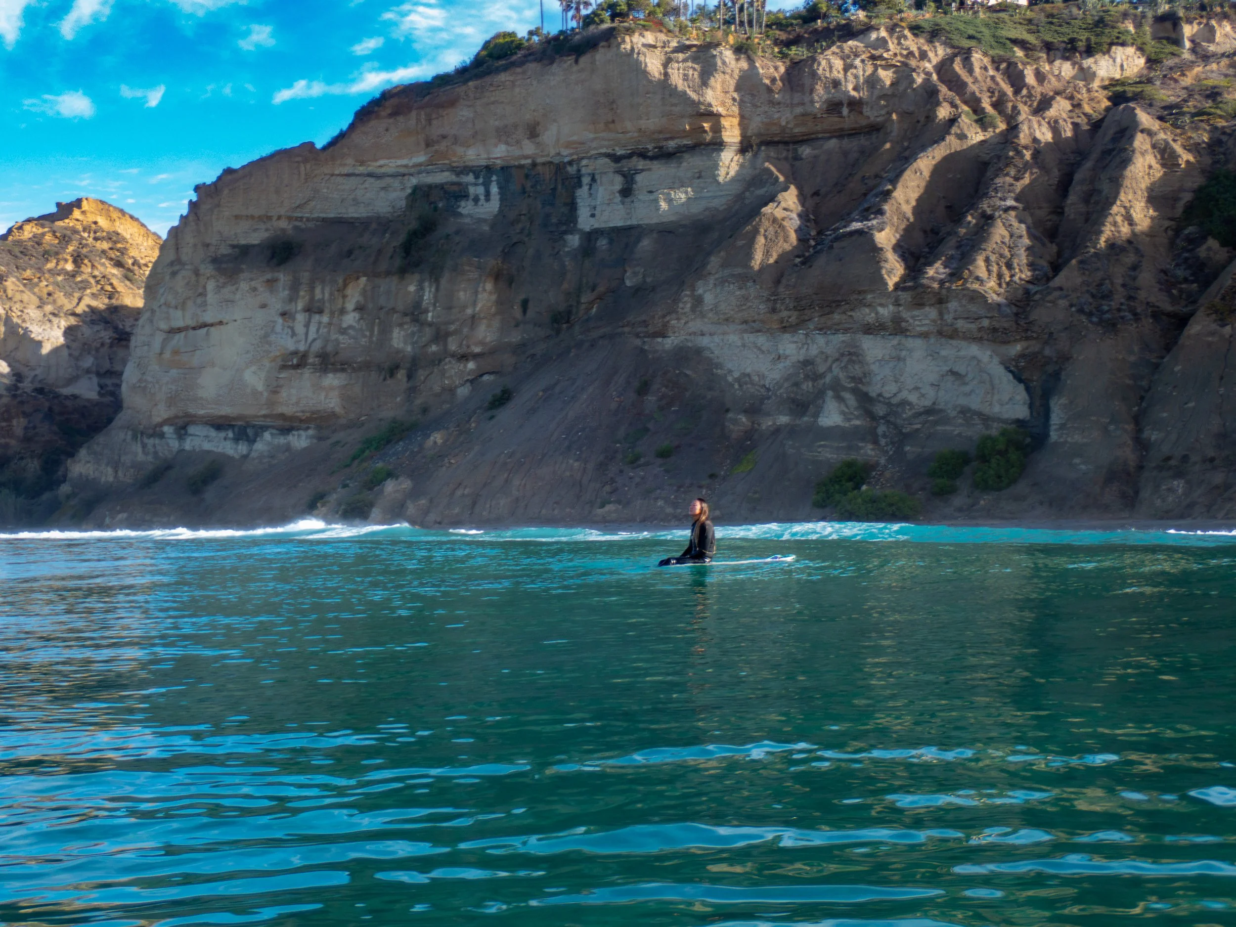Person in the water near a rocky, coastal cliff with trees on top and a partly cloudy sky.