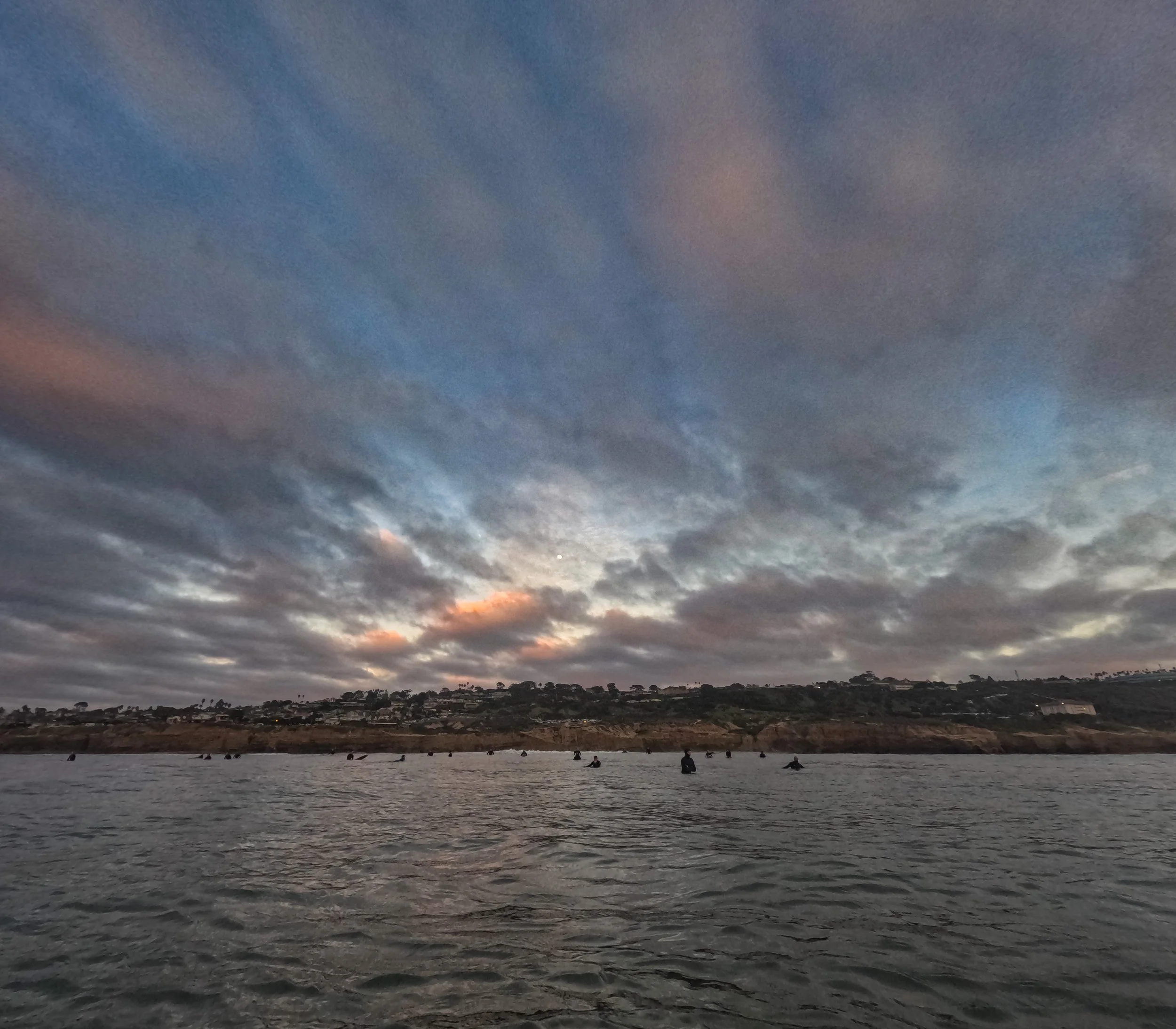View of the ocean at sunset with people in the water and a hilly shoreline in the background under a cloudy sky.
