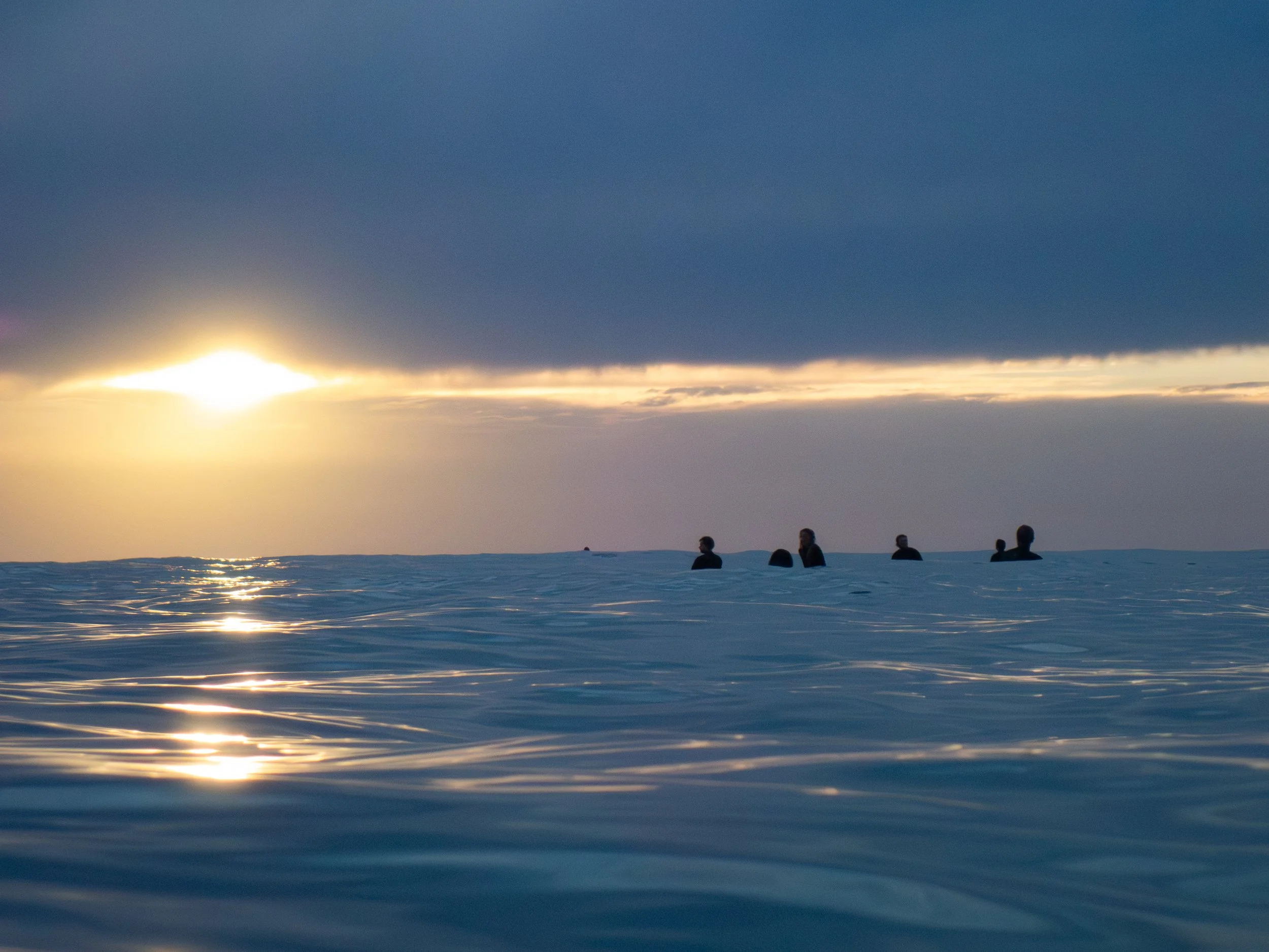 Six people are floating in the ocean during sunset or sunrise, with dark clouds overhead and a bright sun near the horizon.