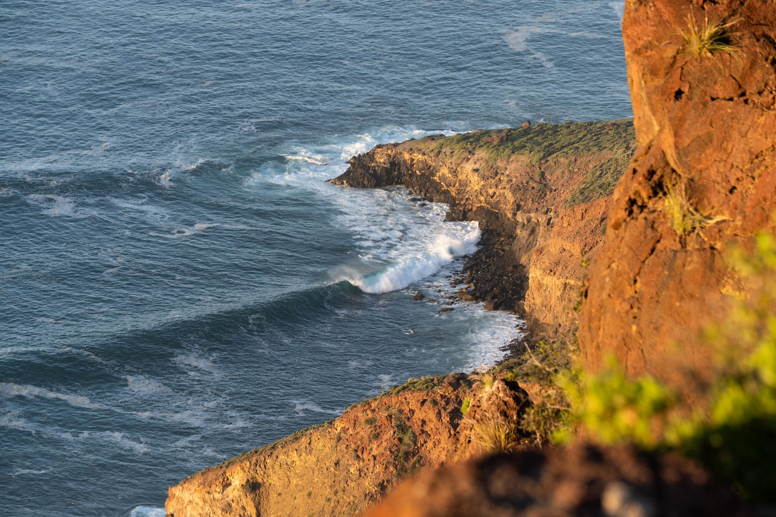 Cliffside overlooking the ocean with waves crashing against rocky shoreline, captured during sunset.
