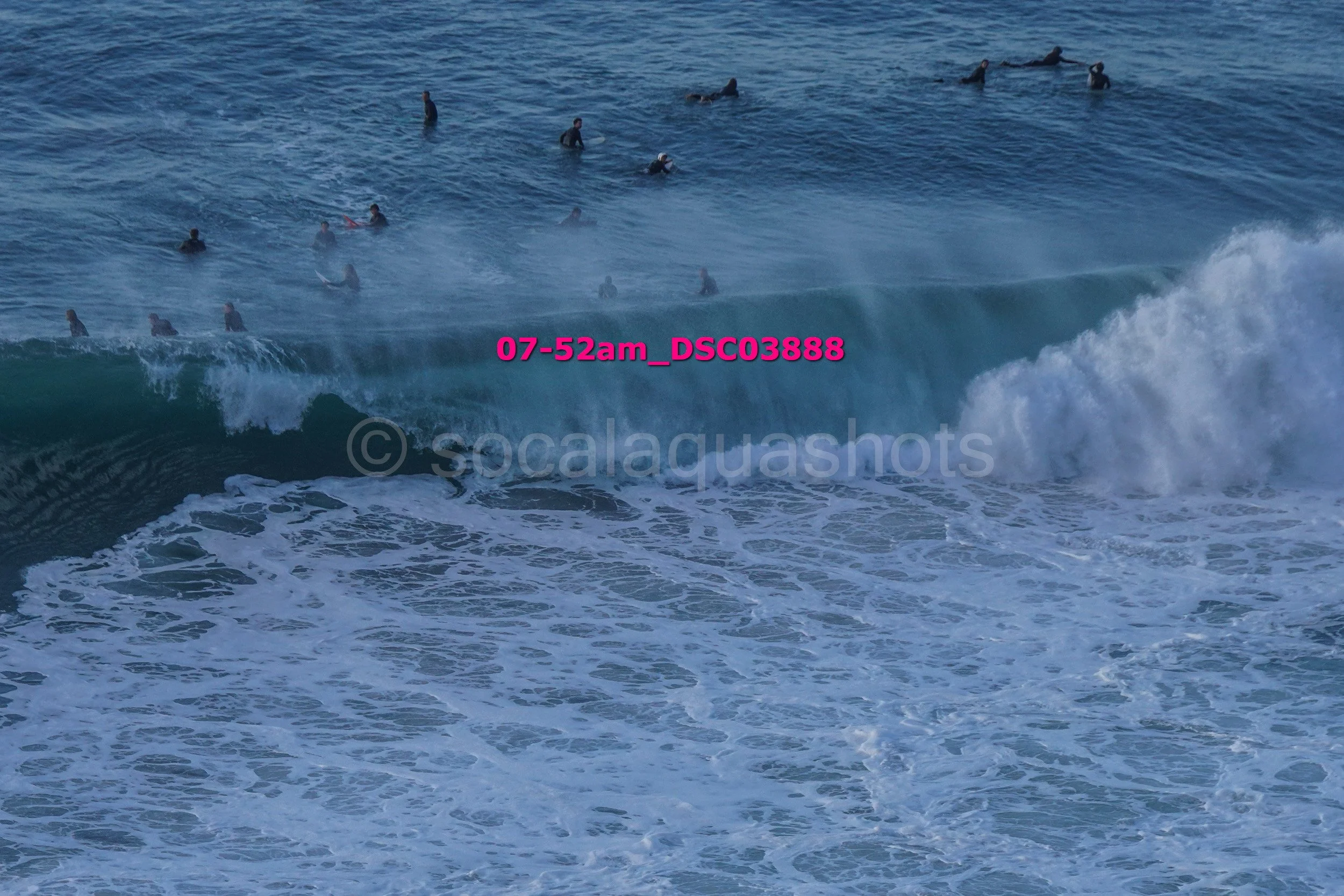 A group of surfers in the ocean, some sitting on their boards, others riding waves, with a large ocean wave in the foreground.