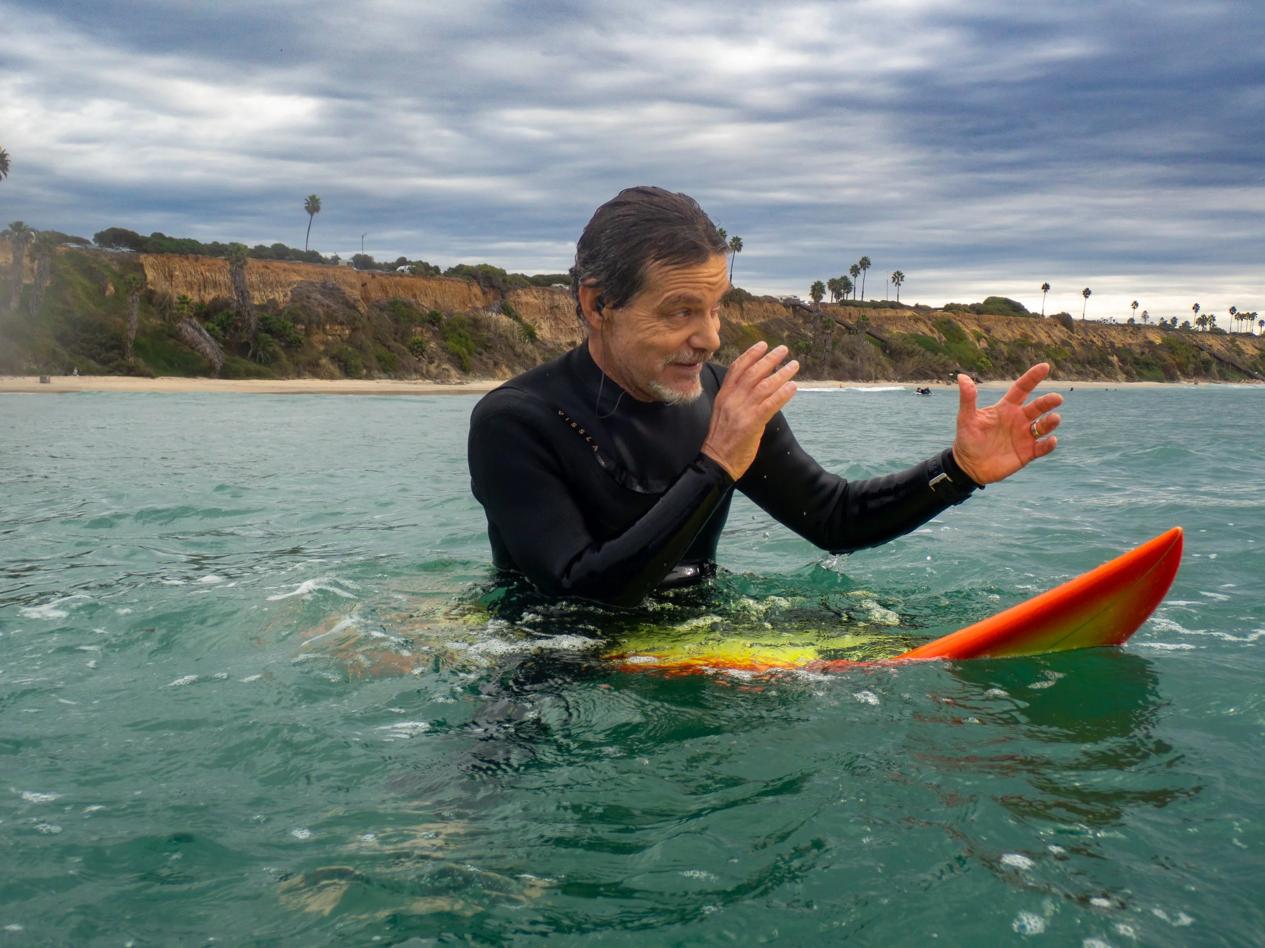 Older man in a wetsuit standing on a surfboard in the ocean, gesturing with his hands, with a coastline and palm trees in the background, under cloudy skies.