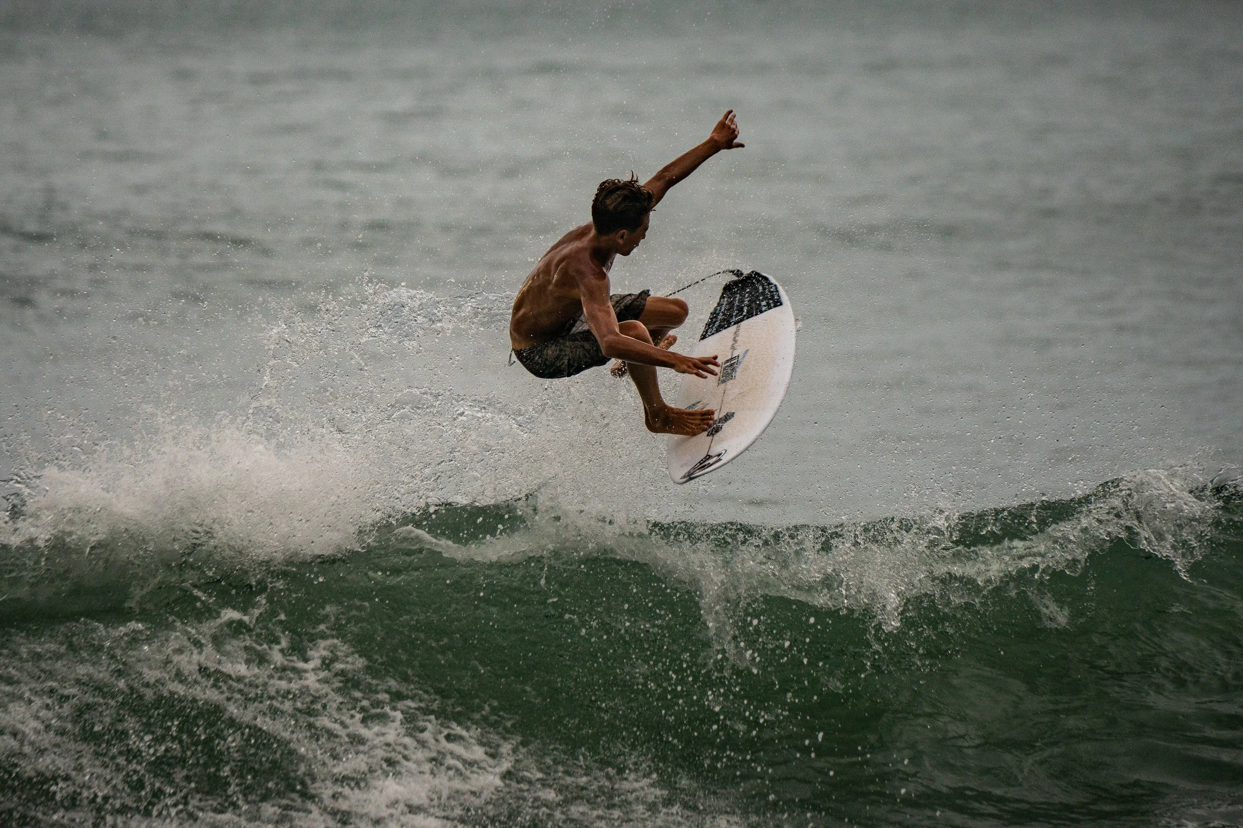 A young man surfing on a wave in the ocean, wearing dark shorts, balancing on a white surfboard with black and gray accents.