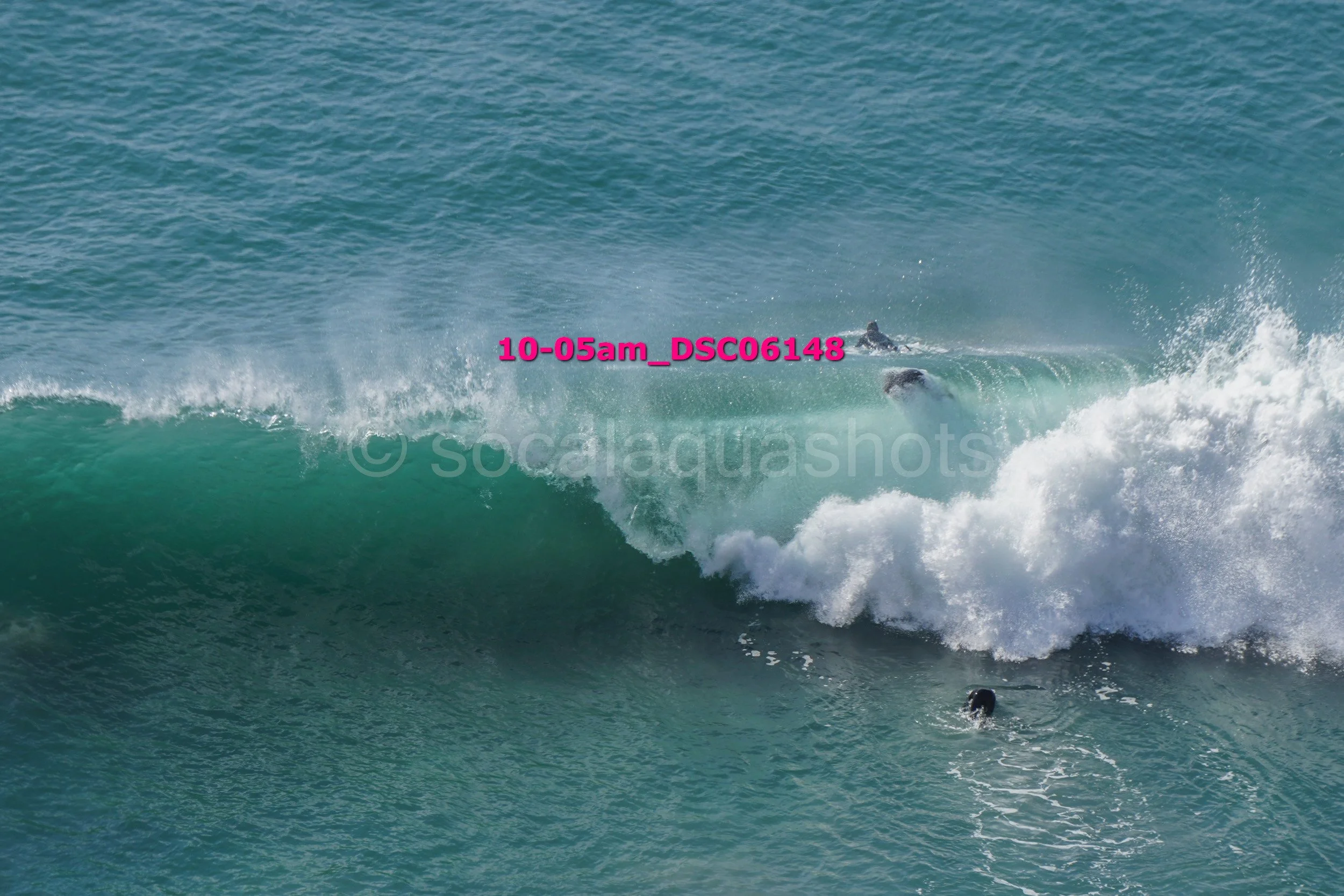 A surfer riding a wave at the beach, with water splashing around them and another surfer swimming nearby.