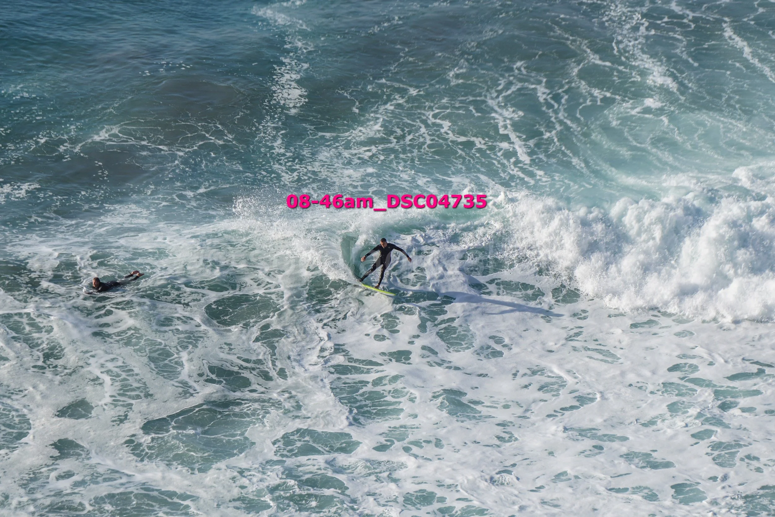 A person surfing on a wave in the ocean, with another swimmer nearby. The water is choppy with white foam, and the scene is bright with sunlight.