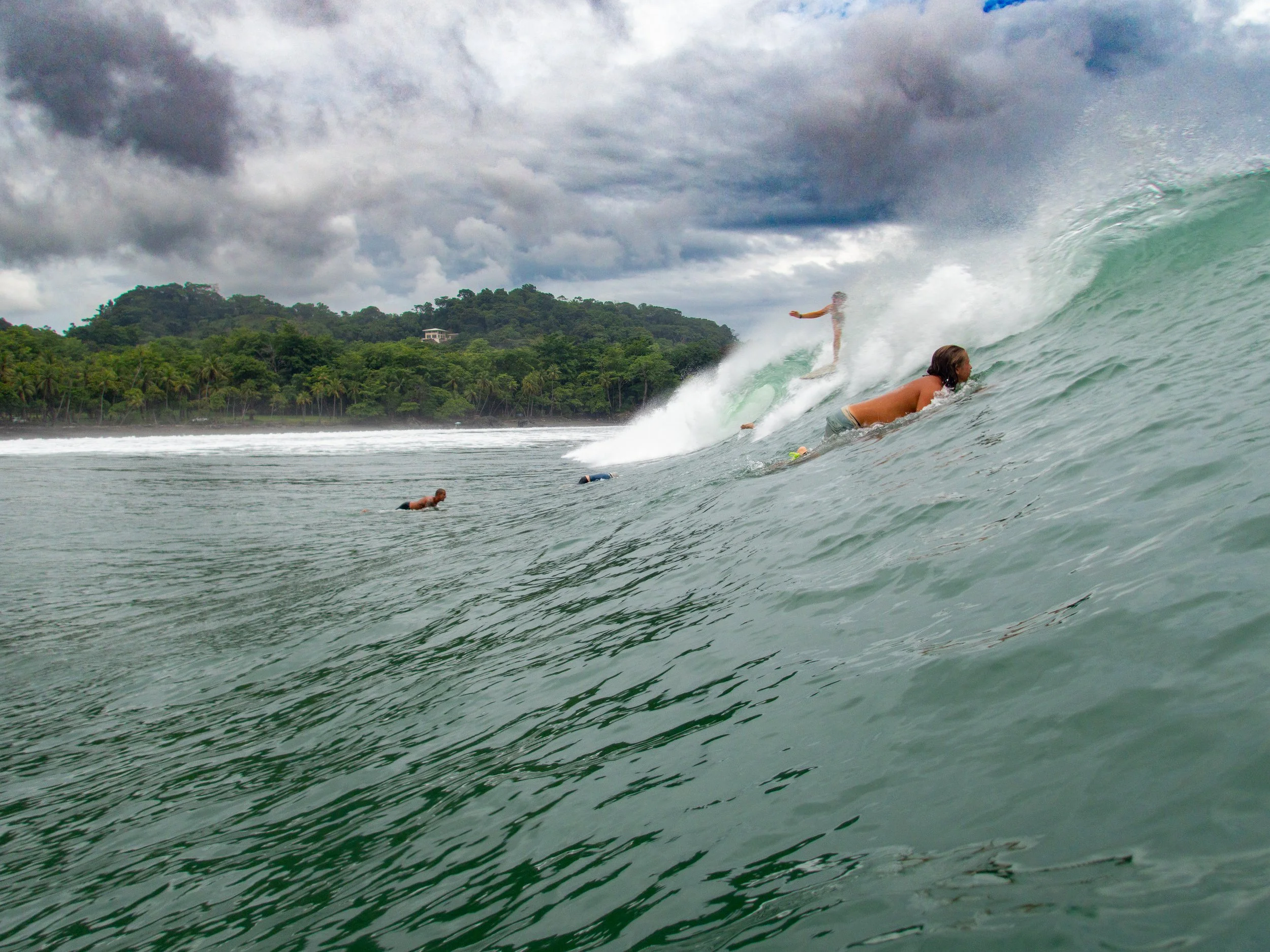 Surfers riding a wave near a tropical coastline with lush green hills under cloudy skies.