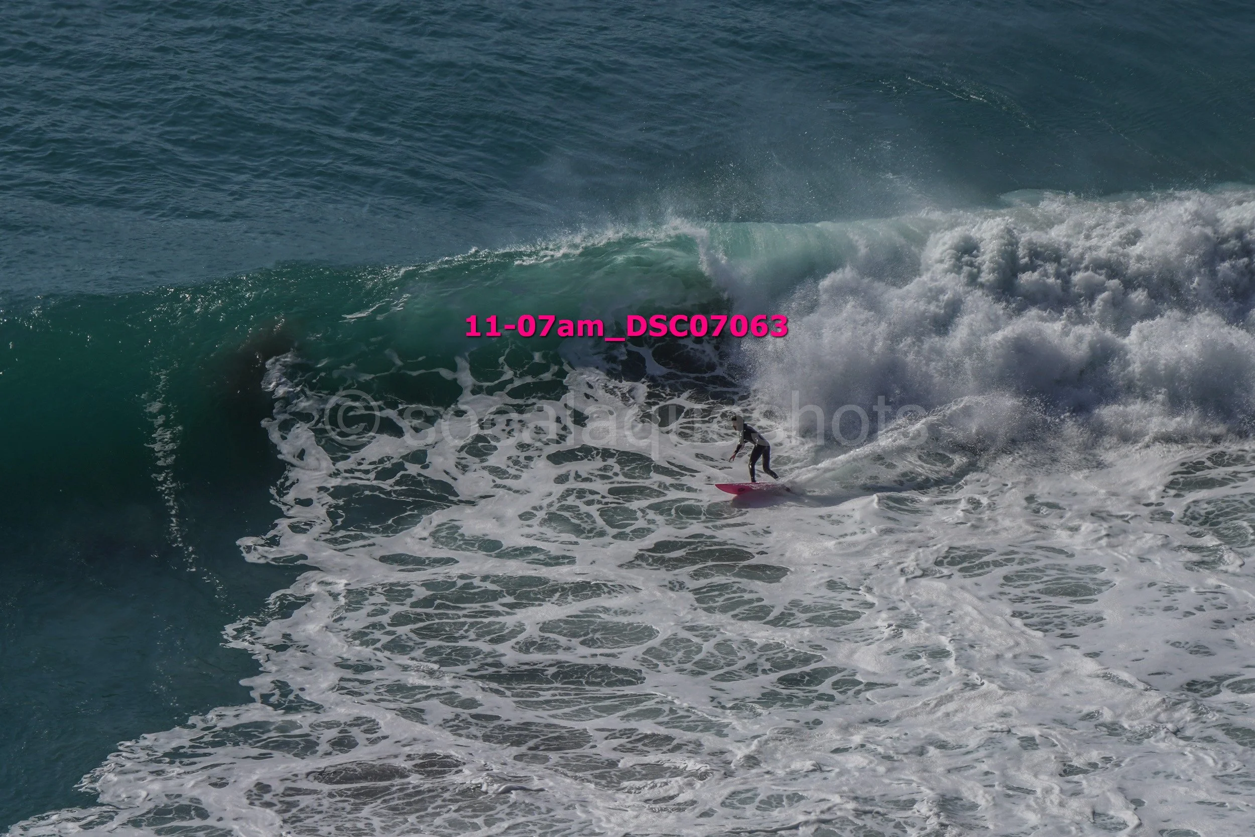 A person surfing on a large wave in the ocean, wearing a wetsuit and riding a pink surfboard.