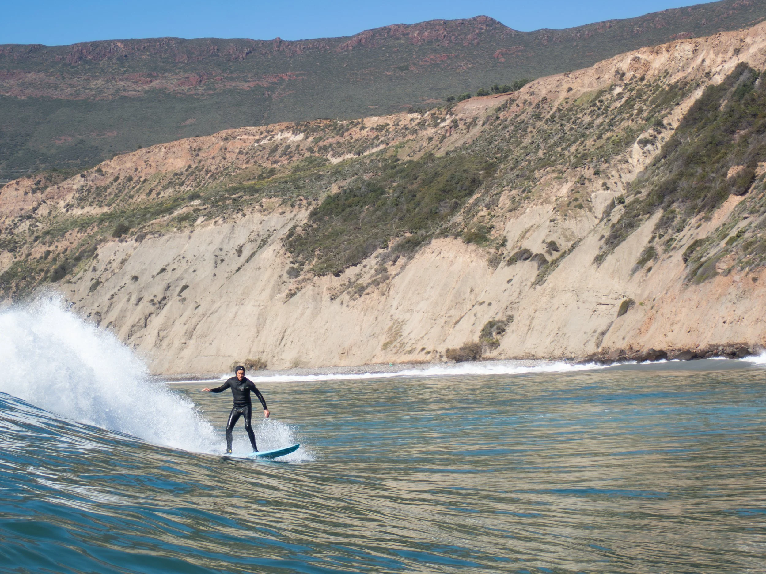 Person surfing on a wave near a rocky beach with cliffs and hills in the background.