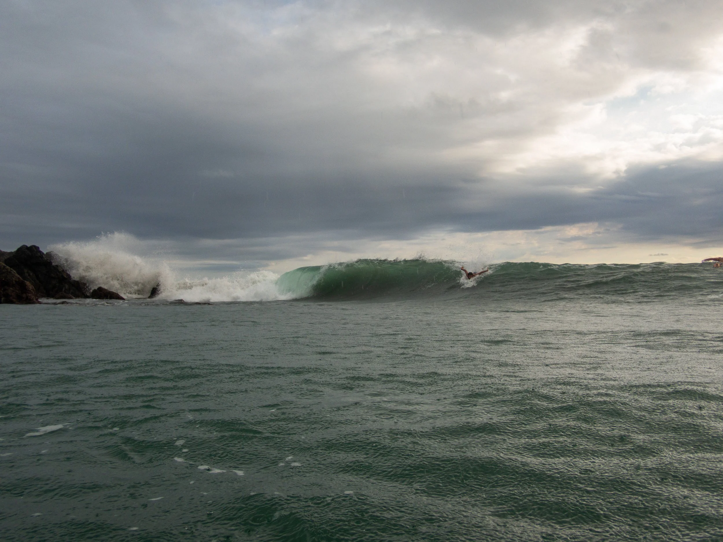 Surfer riding a large wave near rocky shoreline under overcast sky