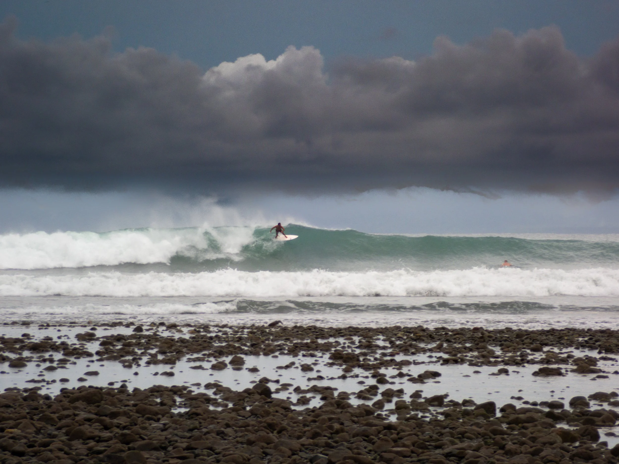 Surfer riding a large wave near a rocky shoreline under a cloudy sky.