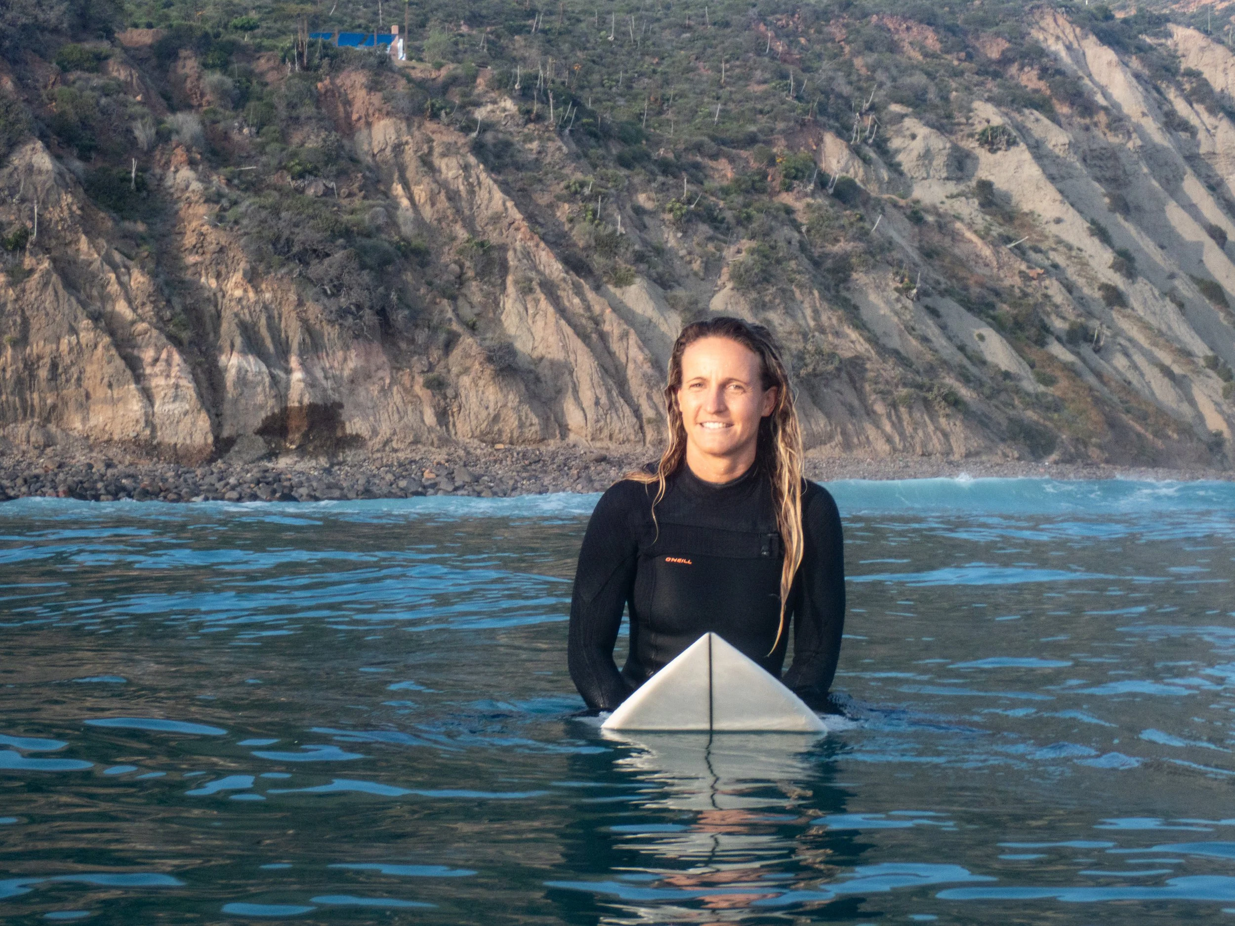 Woman in black wetsuit sitting on surfboard in ocean with rocky cliff and hillside in background
