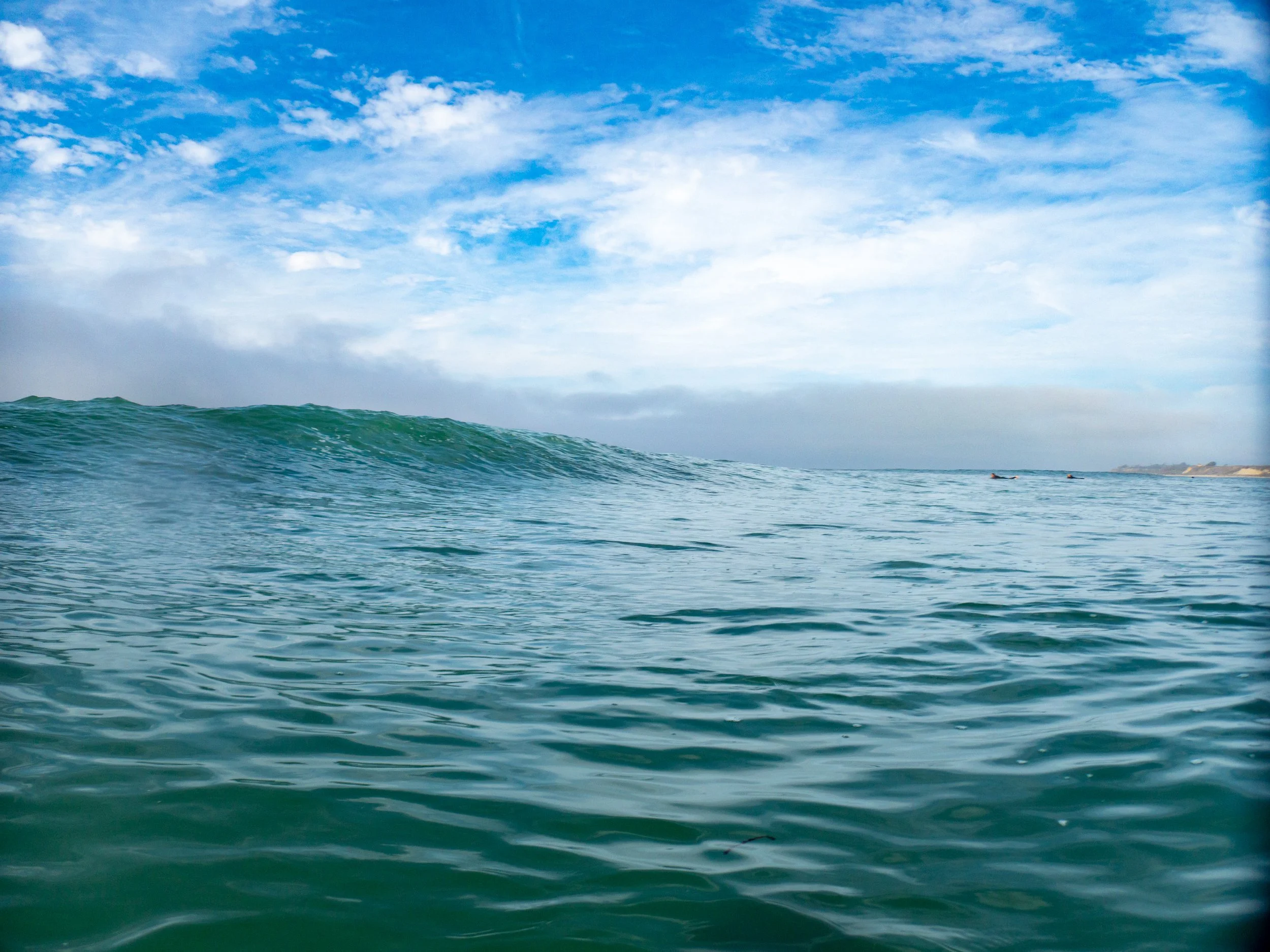 Ocean waves under a partly cloudy sky with some surfers visible in the distance.