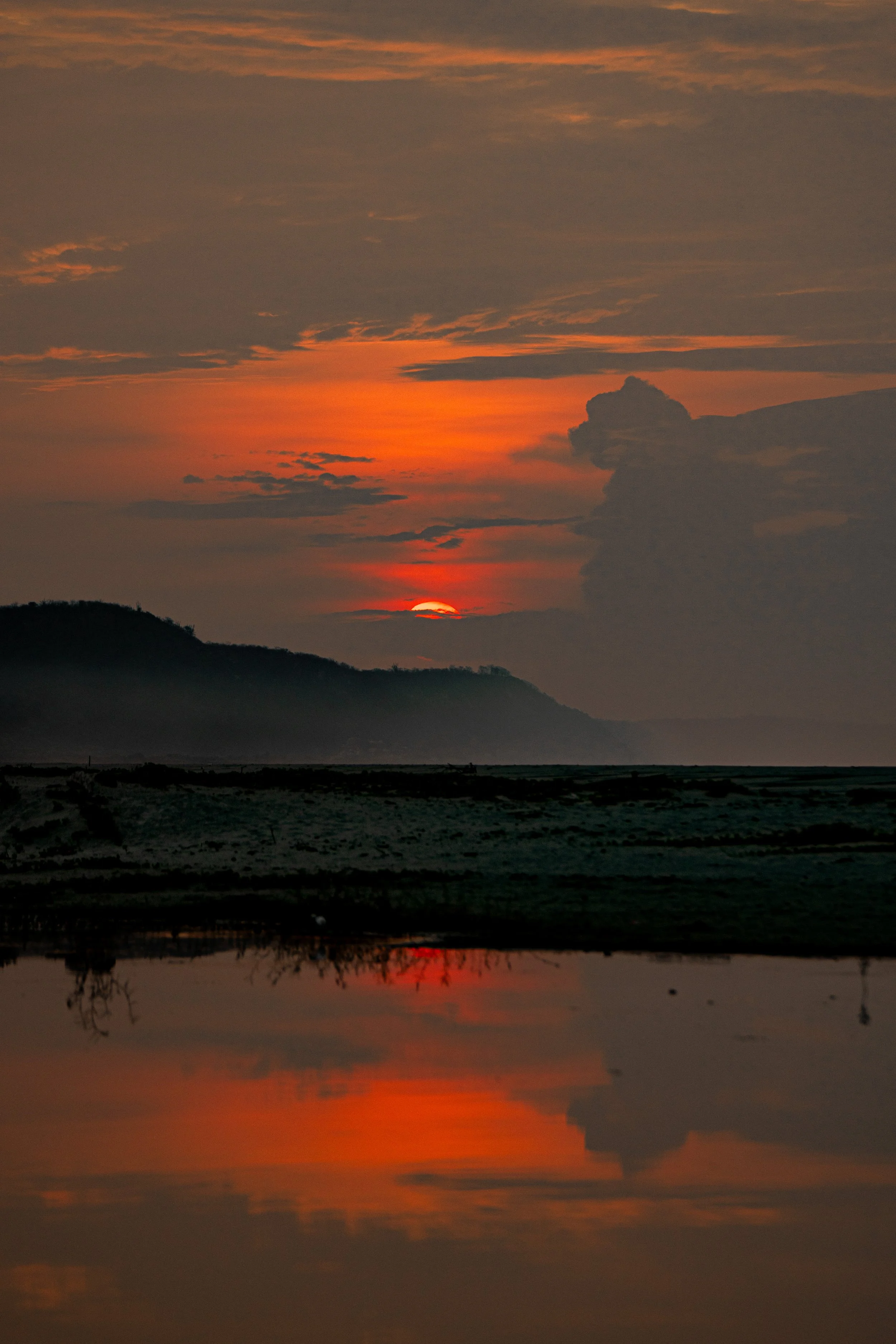 Sunset over a landscape with hills and a body of water, showing a colorful sky with orange, red, and gray clouds.