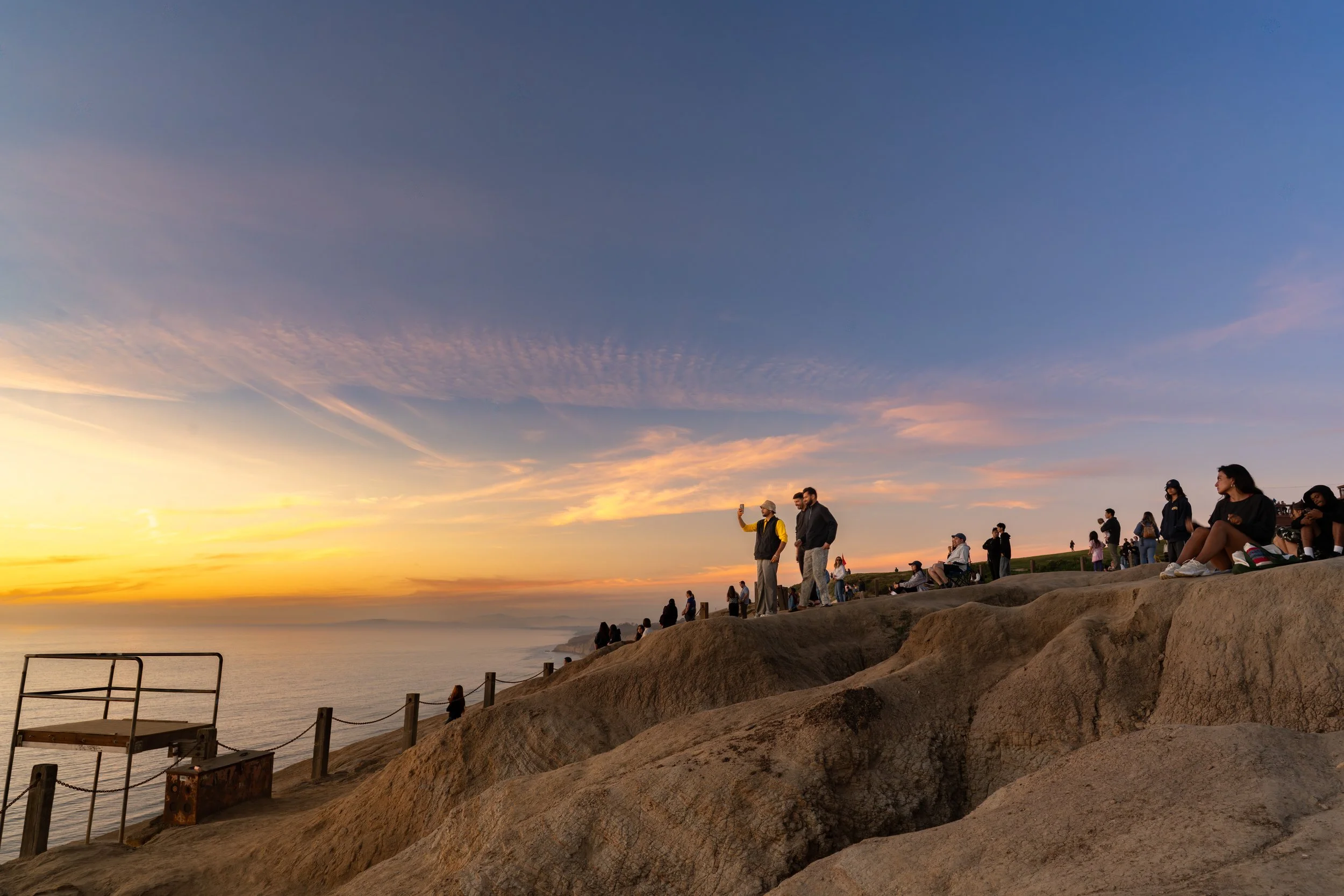 People sitting and standing on rocky cliffs watching a sunset over the ocean.