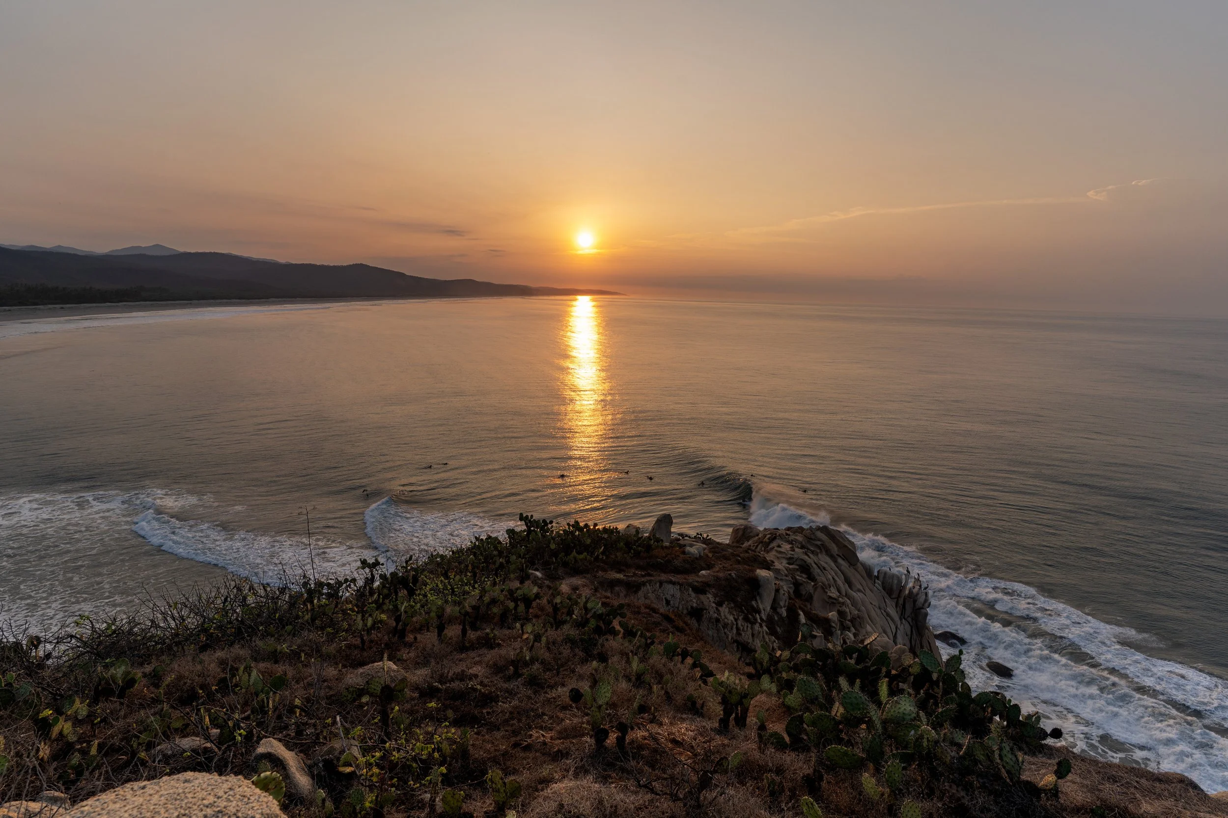 Sunset over the ocean with calm waves, coastal rocks in the foreground, and distant mountains on the horizon.
