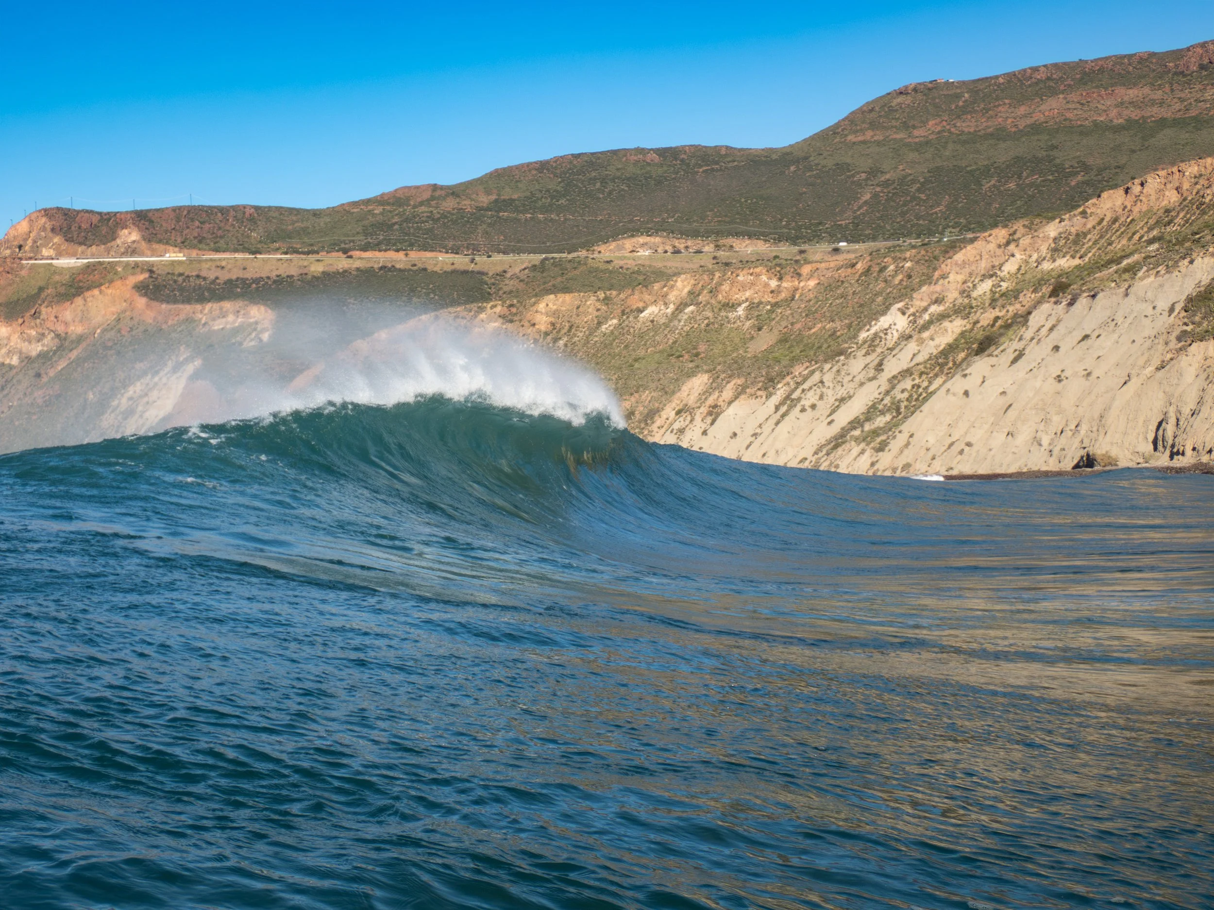 Ocean wave near coastal cliffs with green hills in the background under a clear blue sky.