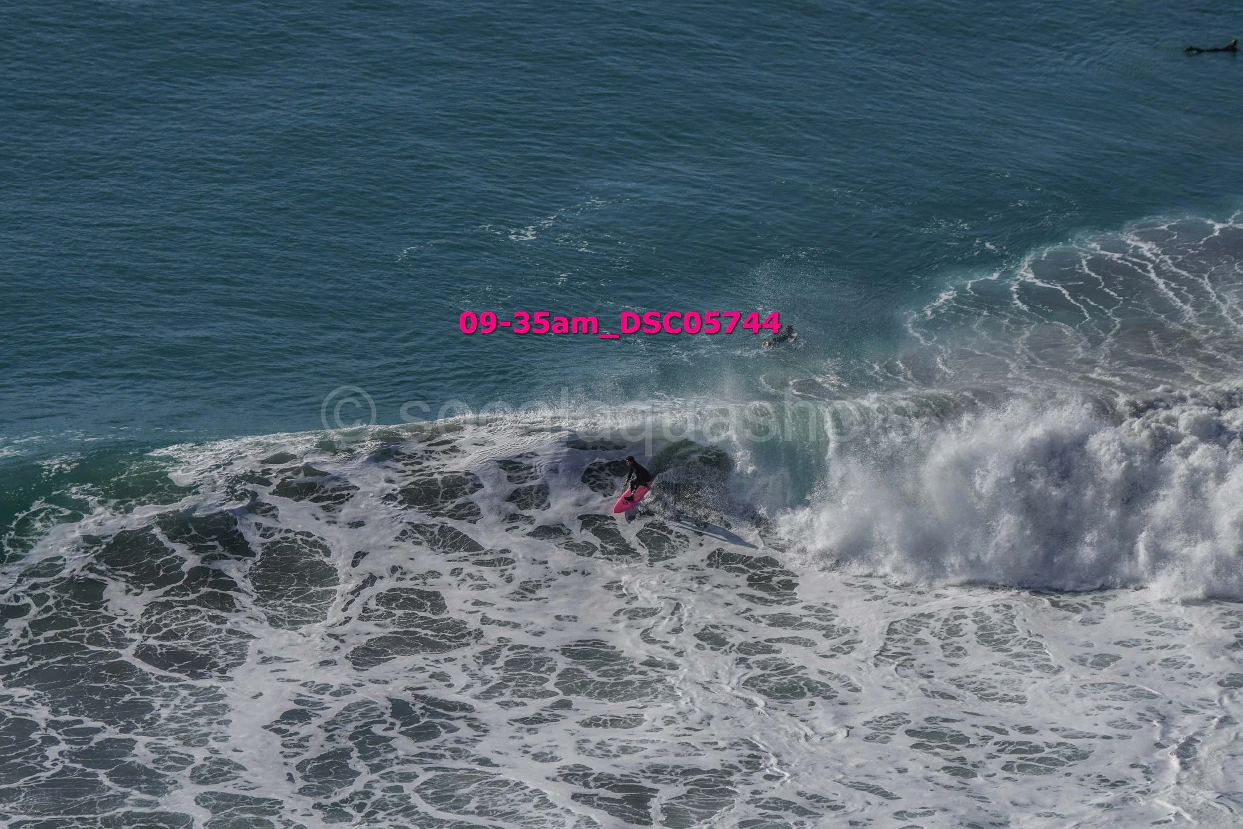 A person surfing on a pink surfboard in the ocean amidst waves, with the sky visible in the background and a few other surfers in the distance.