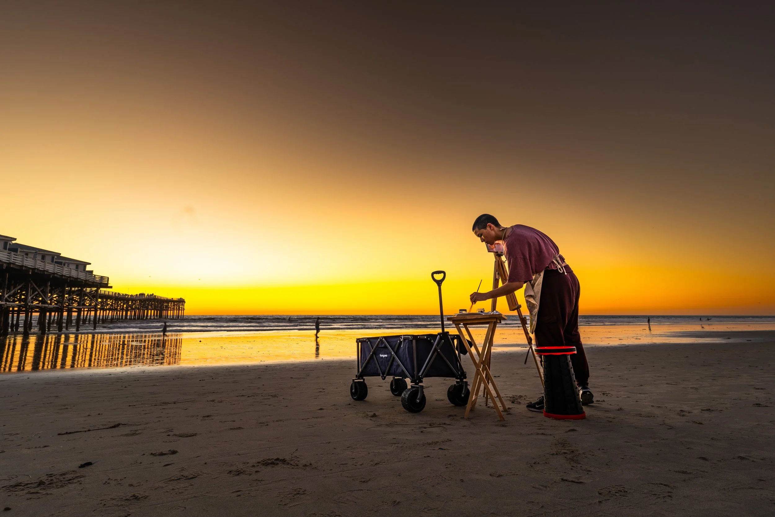 A man on the beach at sunset, setting up equipment with a shovel and a wheeled cart, near a pylon and a pier extending into the water.