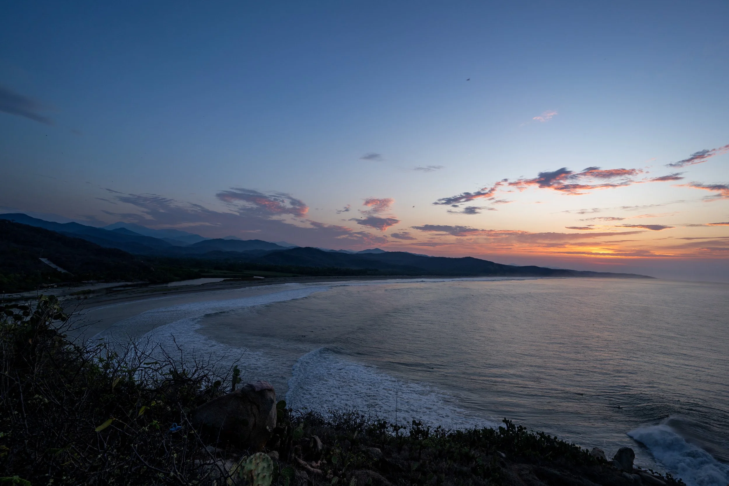 Sunset over a coastal landscape with hills in the background and waves crashing onto the beach.