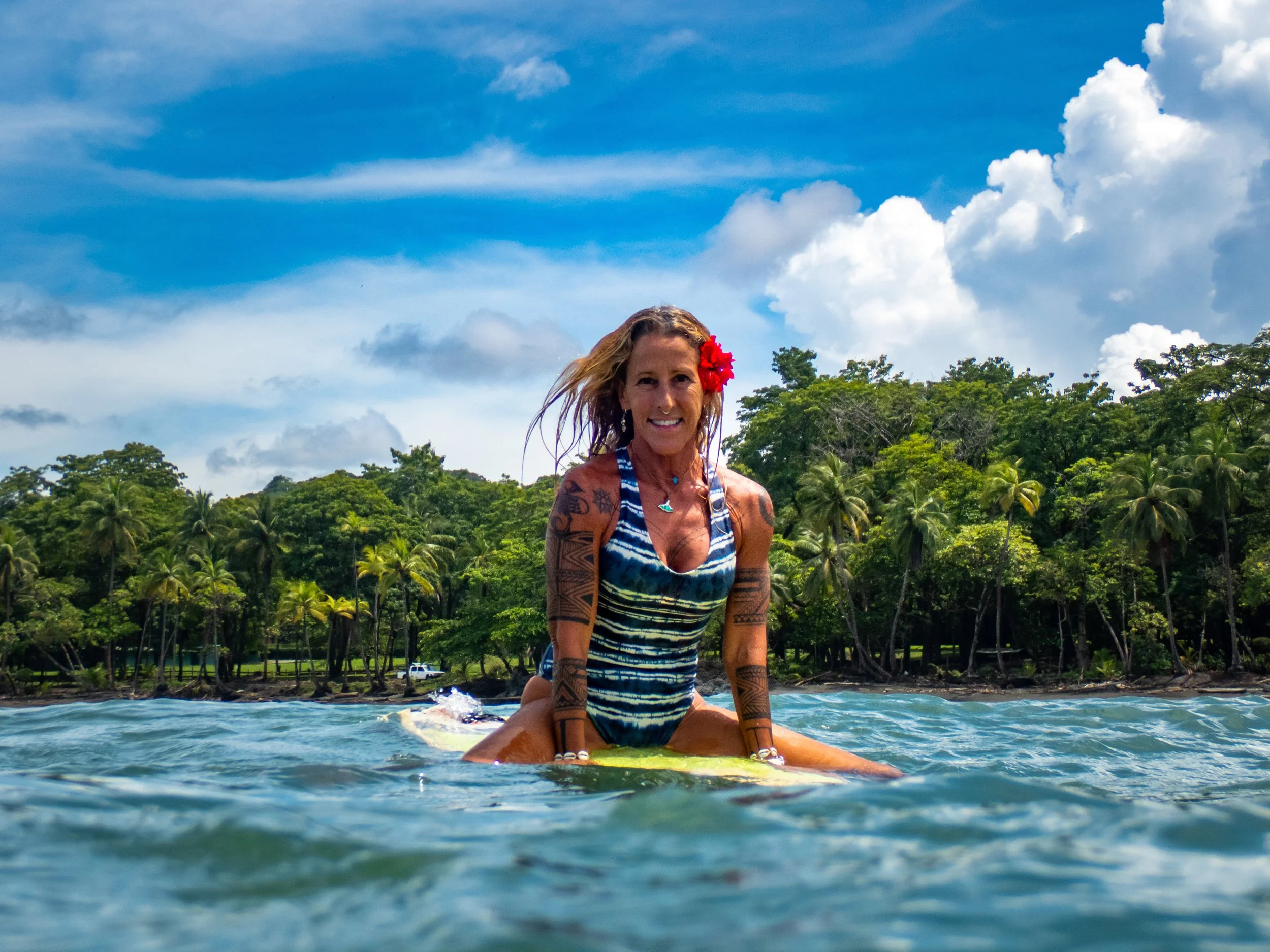 A woman with tattoos, wearing a blue striped swimsuit and a red flower in her hair, surfing on a yellow surfboard in the ocean with a background of green trees and a partly cloudy sky.