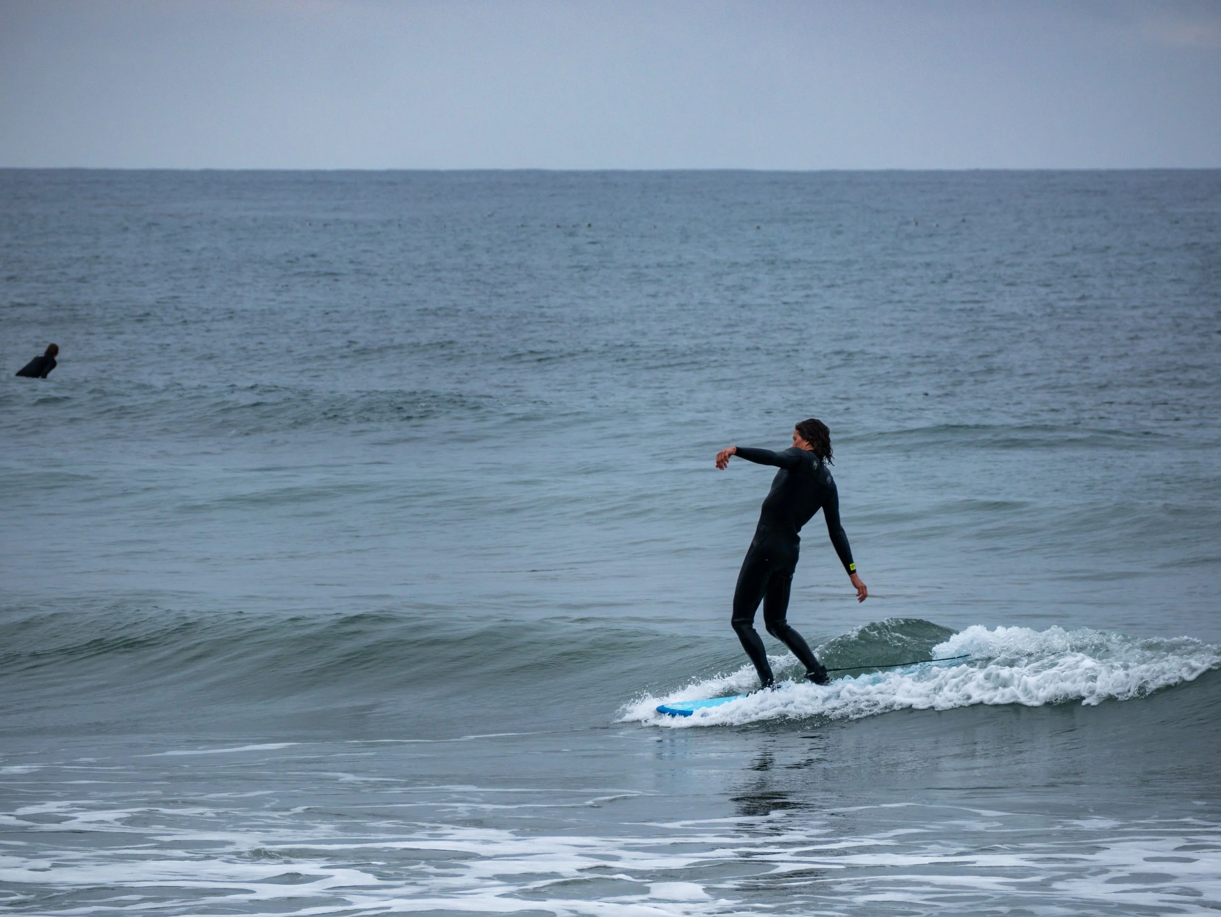 A person in a wetsuit riding a small wave on a surfboard at the beach, with another person in the water in the background.