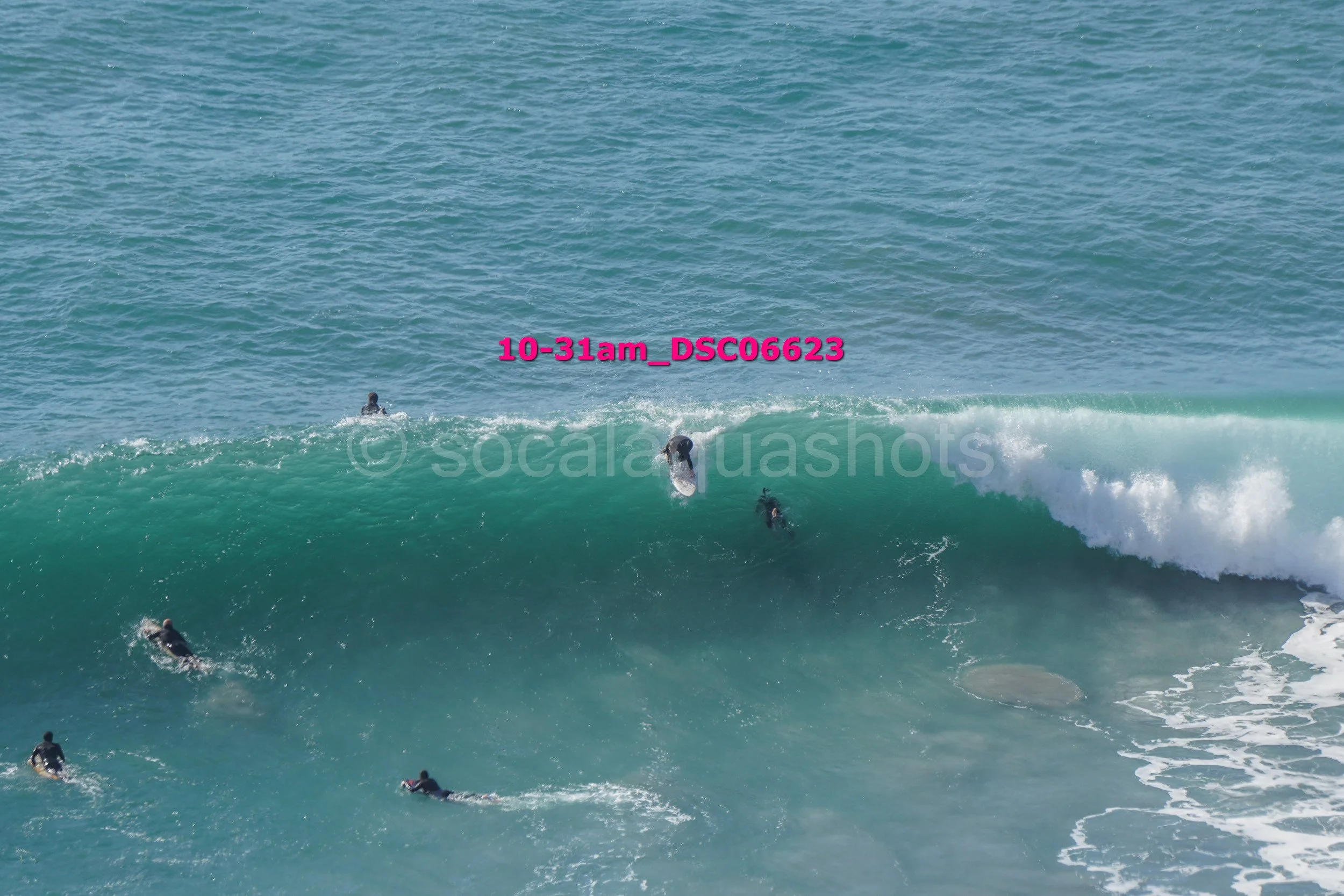 Surfers riding a wave in the ocean, with multiple surfers visible across the water.