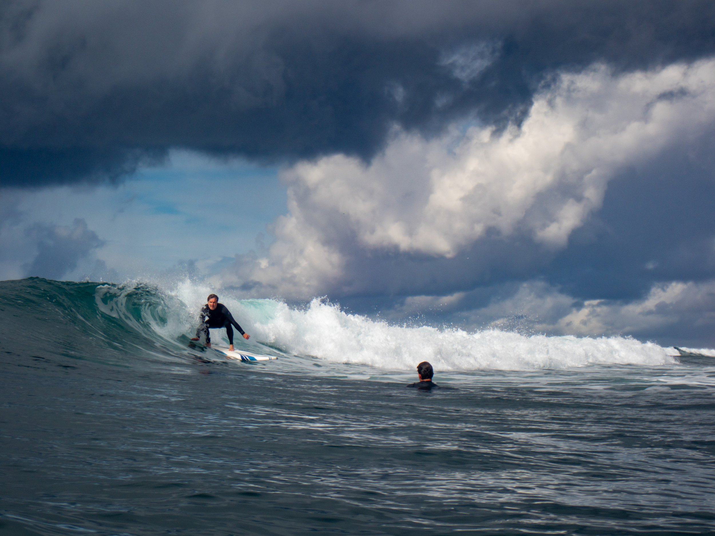 A person surfing on a large wave with dark stormy clouds in the sky and a person swimming in the water nearby.