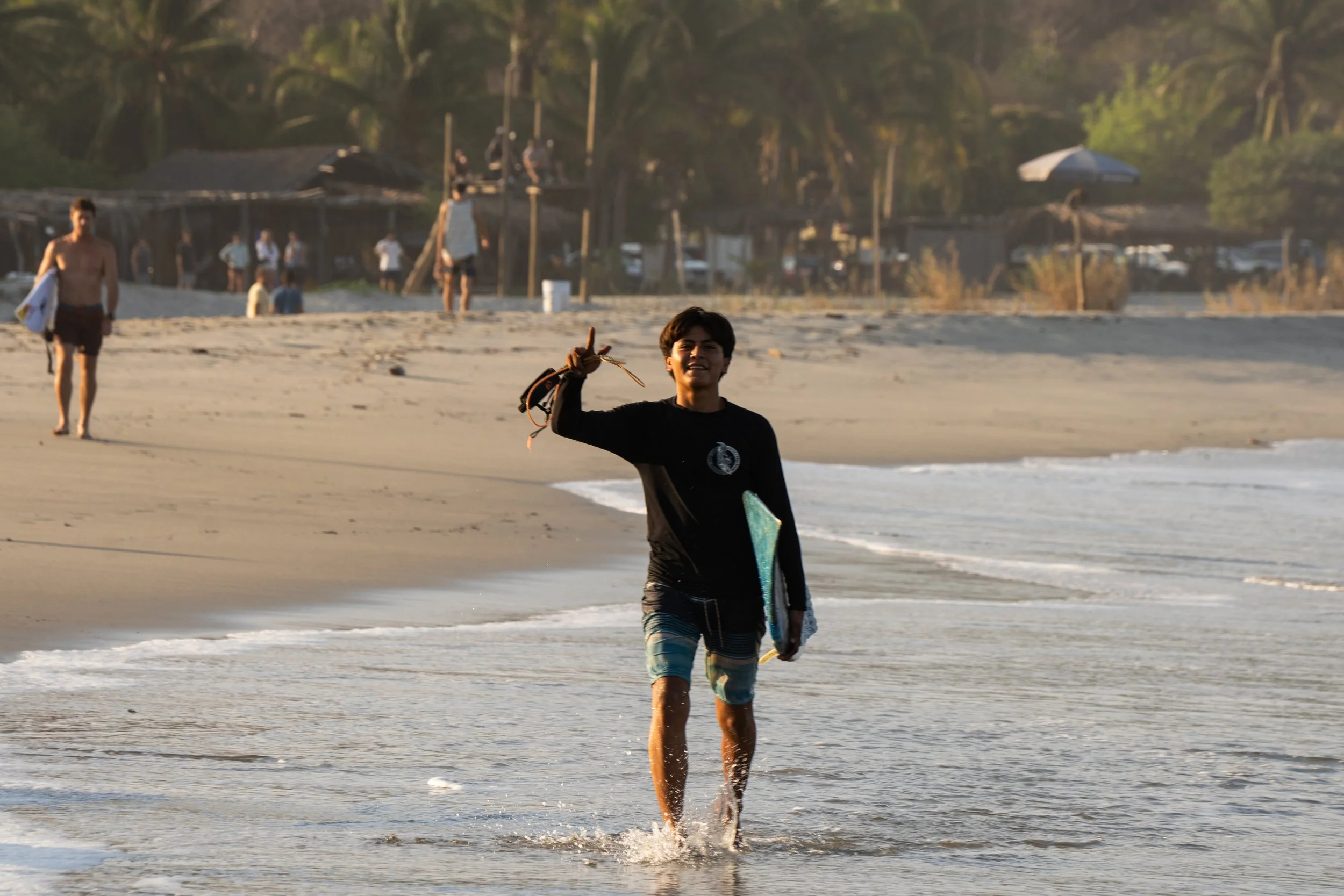A young boy walking out of the ocean with a surfboard in one hand and a towel in the other, smiling, with people and trees in the background on a beach.